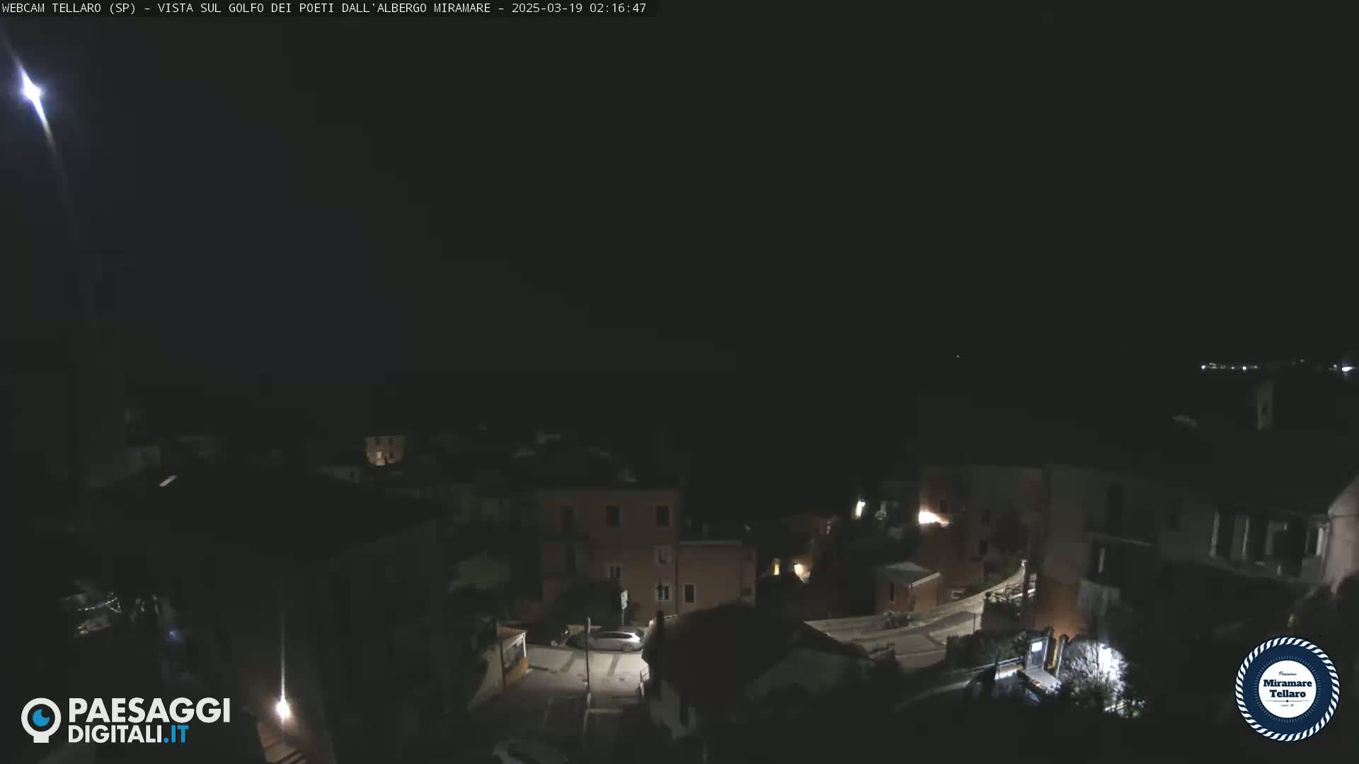 A nighttime view of a small town with a few lit buildings and a single car, under a dark, clear sky with a bright light possibly a star or plane in the upper left corner.