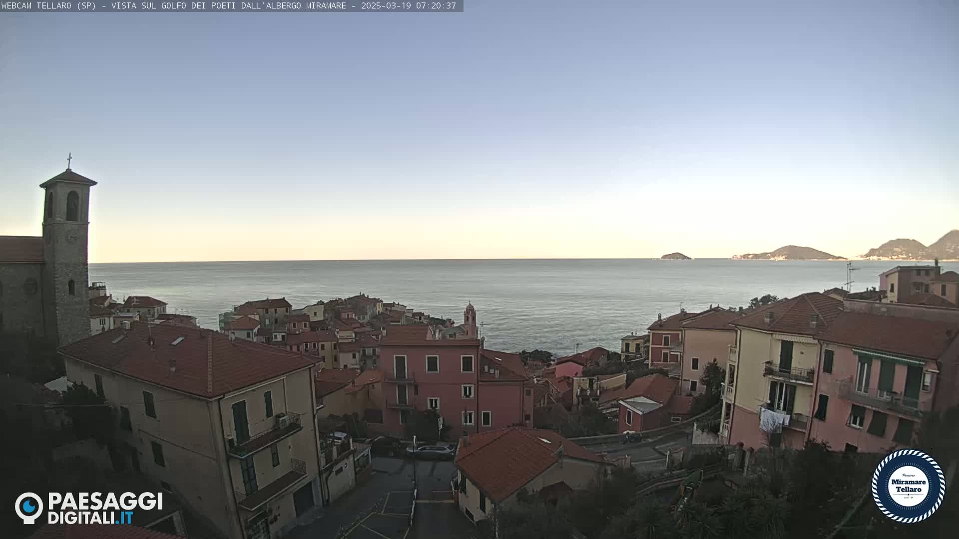 A coastal town with red-tiled roofs and a church steeple overlooks a calm sea under a clear, light-blue sky.