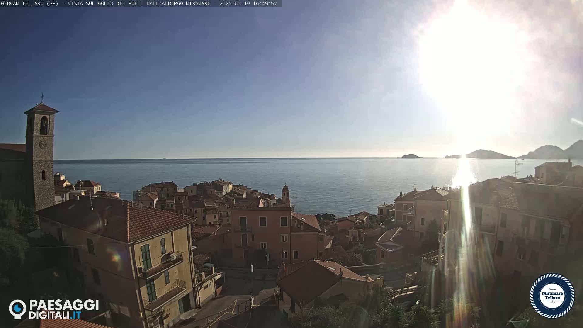 A coastal town with red-tiled roofs and a bell tower overlooks a calm sea under a bright, sunny sky.