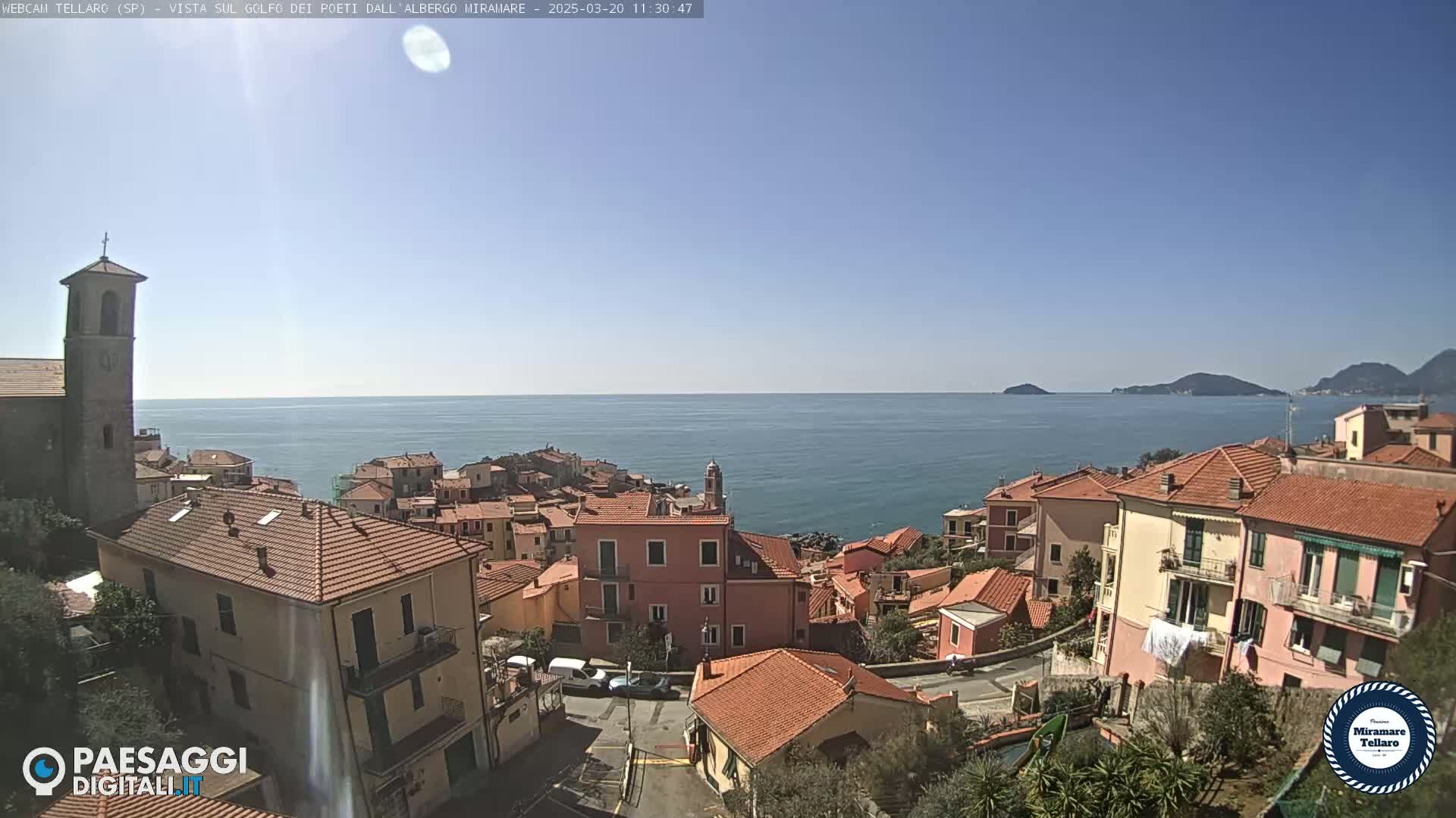 A coastal town with red-tiled roofs, a church steeple, and a calm blue sea under a clear sky is shown.