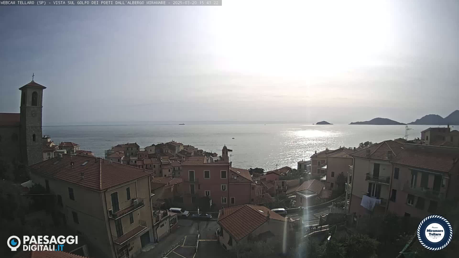 A coastal town with red-tiled roofs overlooks a calm sea under a bright, mostly clear sky.
