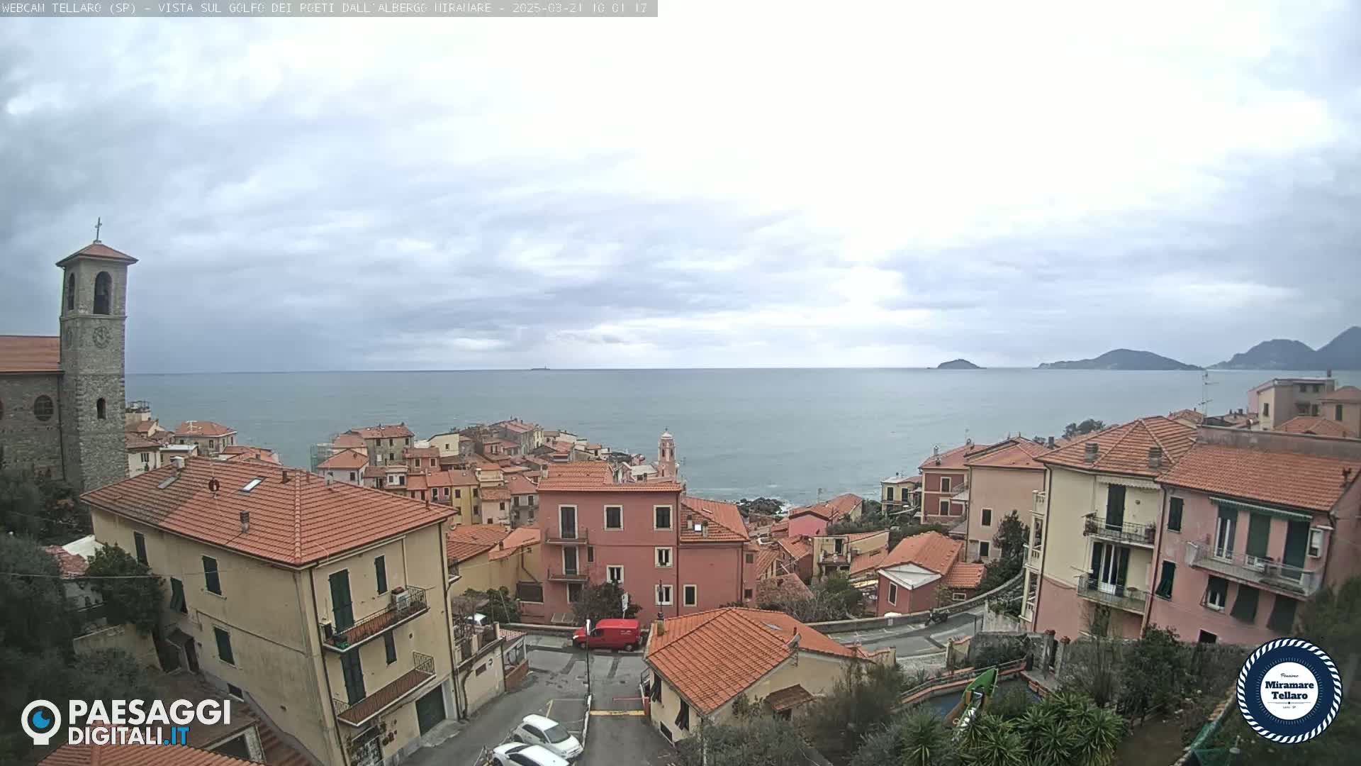 A coastal town with red-roofed buildings overlooks a calm sea under a cloudy sky.