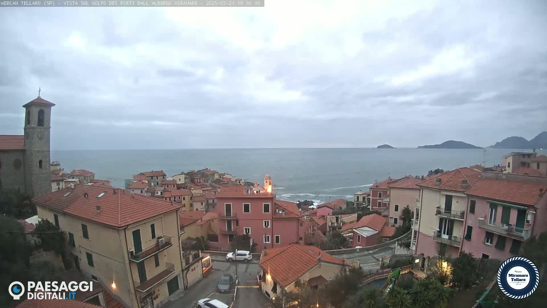 A coastal town with red-tiled roofs and a church overlooks a calm sea under a cloudy sky.