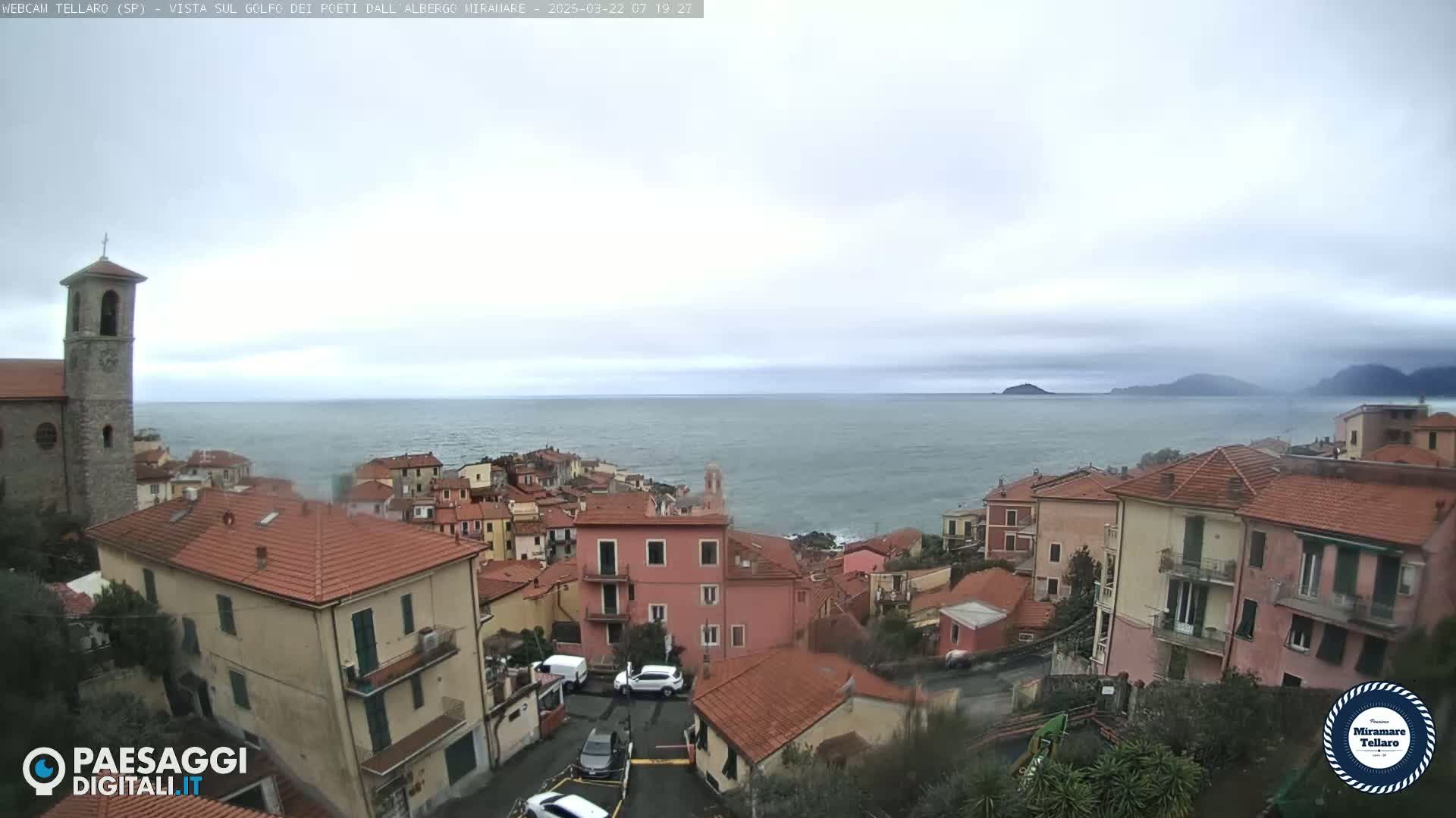A coastal town with red-tiled roofs and a church overlooks a calm sea under an overcast sky.