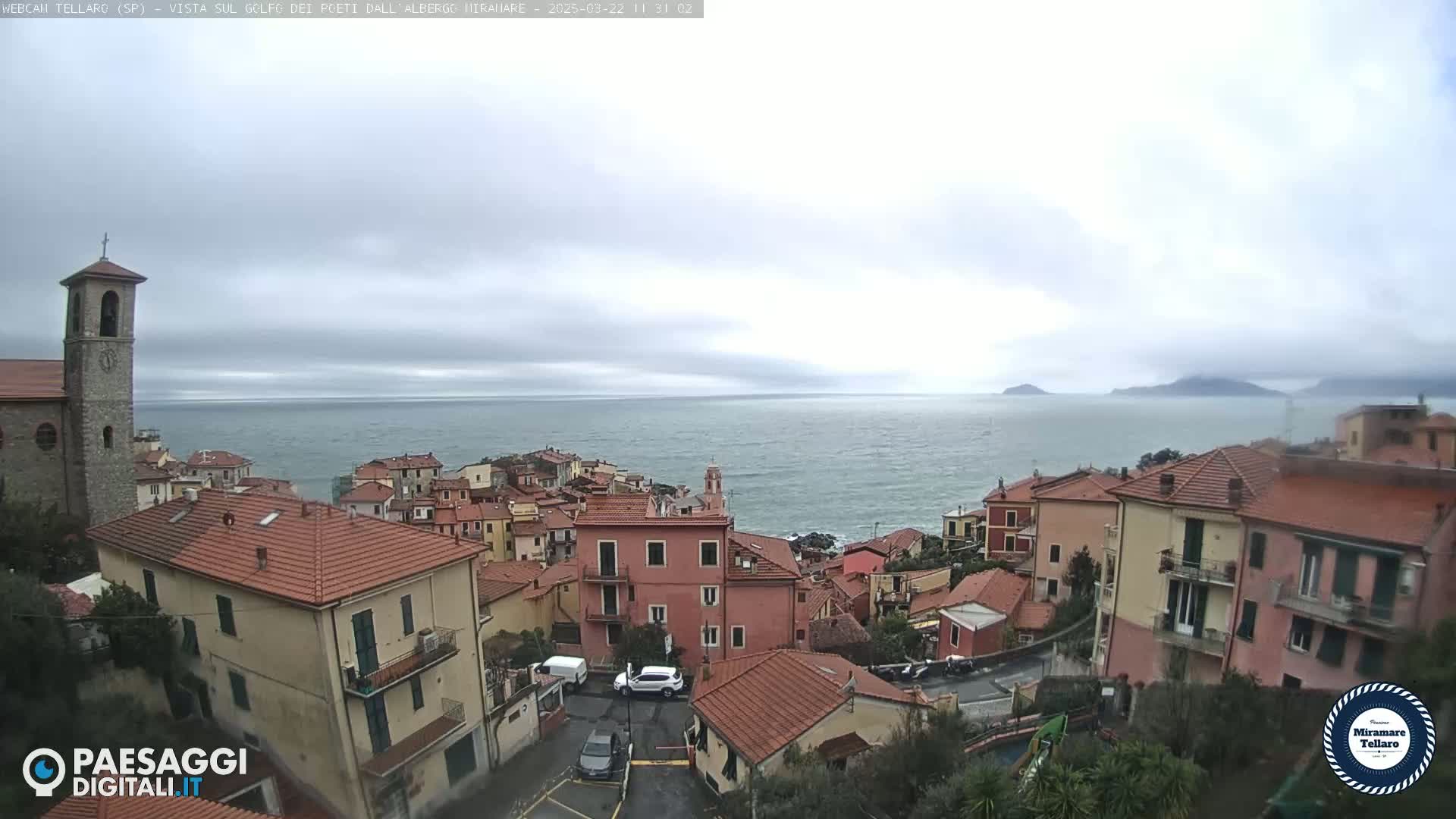 A coastal town with red-tiled roofs and a church steeple overlooks a calm sea under a cloudy sky.