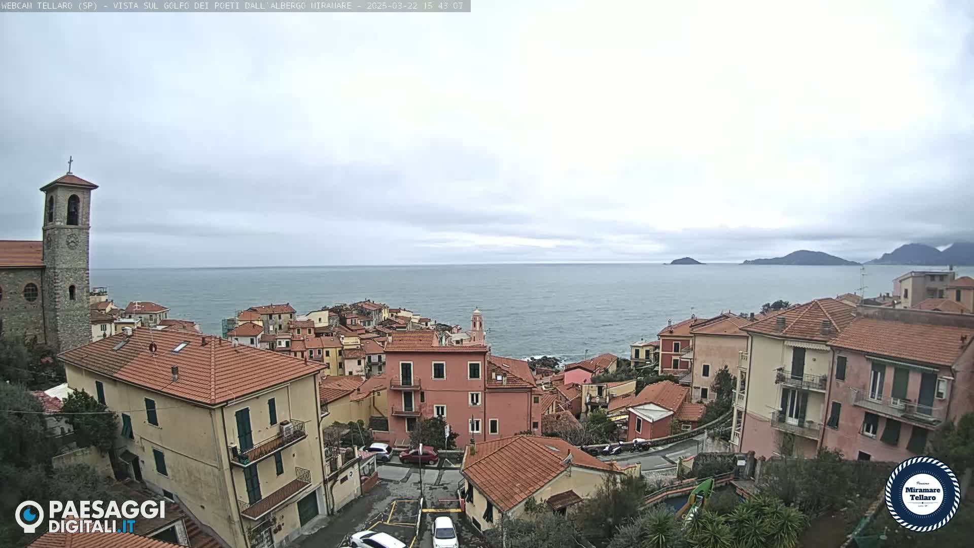 An overcast day reveals a coastal town with red-roofed buildings, a bell tower, and a calm sea with distant islands.