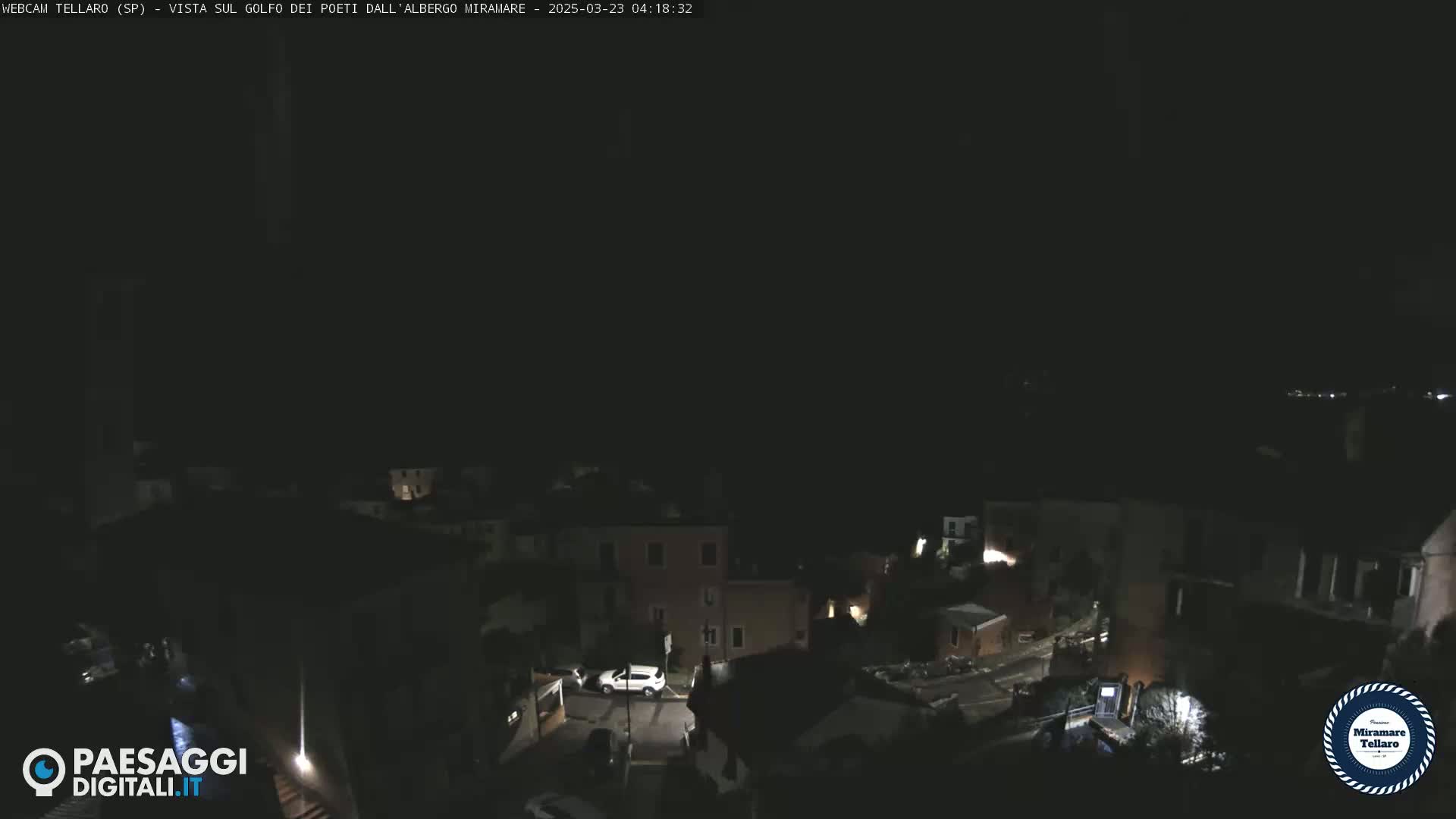 A nighttime, high-angle view of a coastal town with dimly lit buildings and a few cars parked on a street.