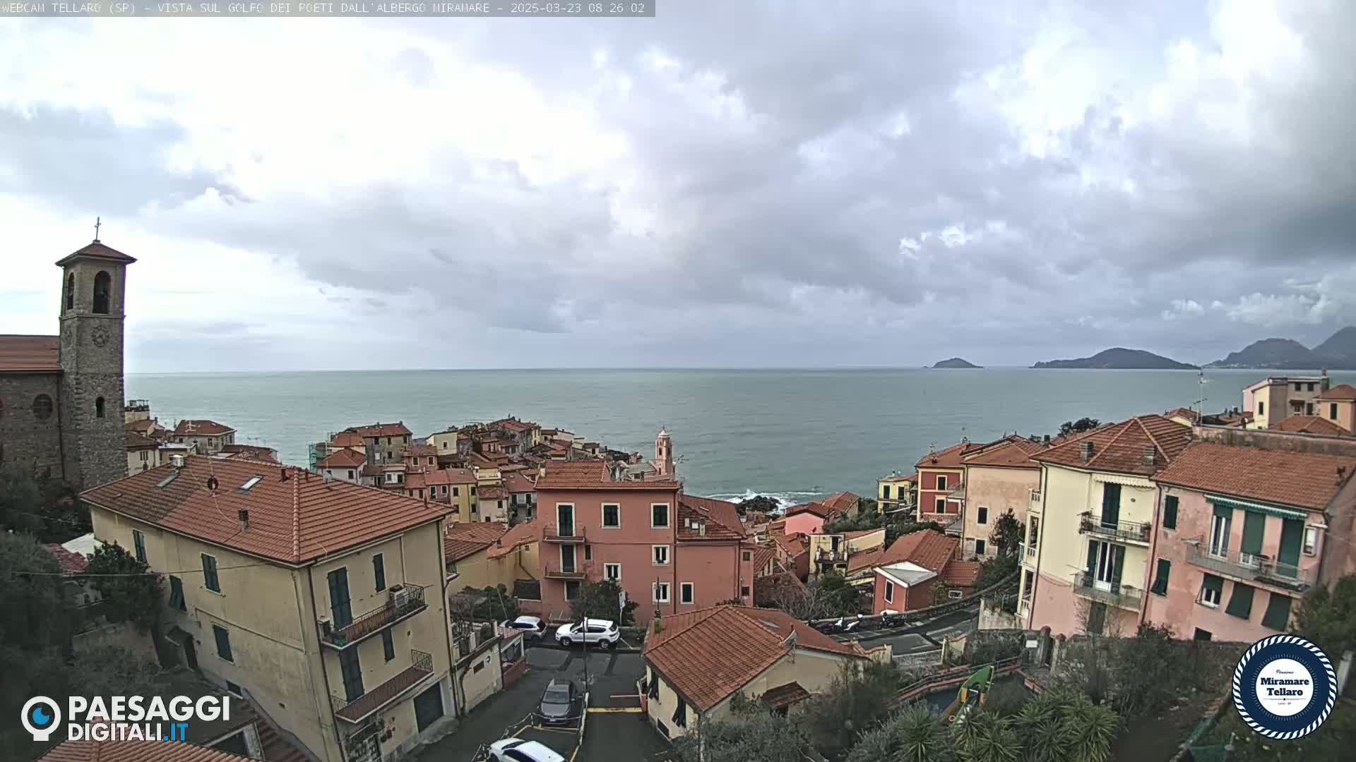 A coastal town with red-tiled roofs and a church steeple overlooks a calm sea under a cloudy sky.