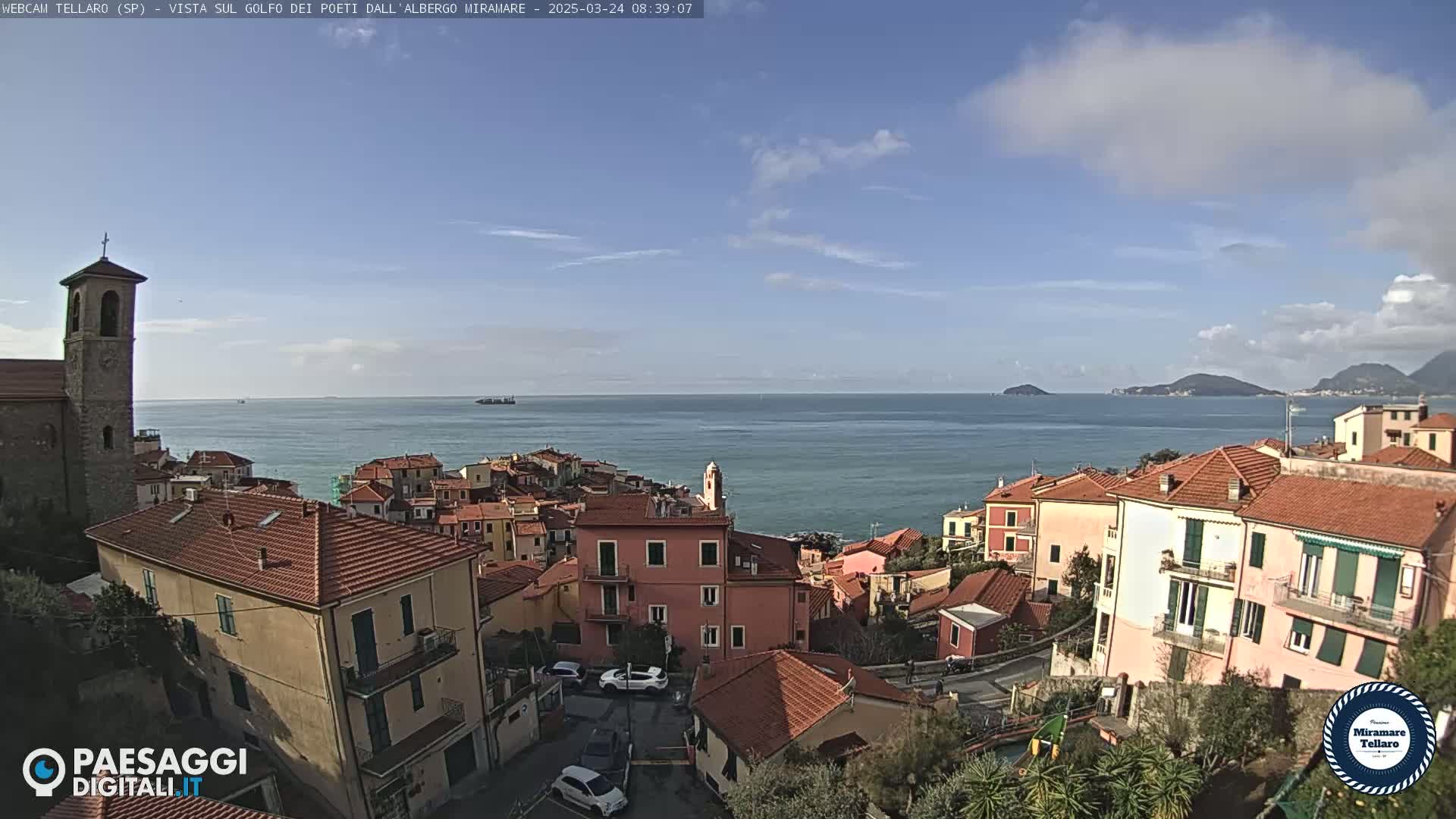 A coastal town with red-roofed buildings overlooks a calm sea under a mostly clear sky with a few scattered clouds.