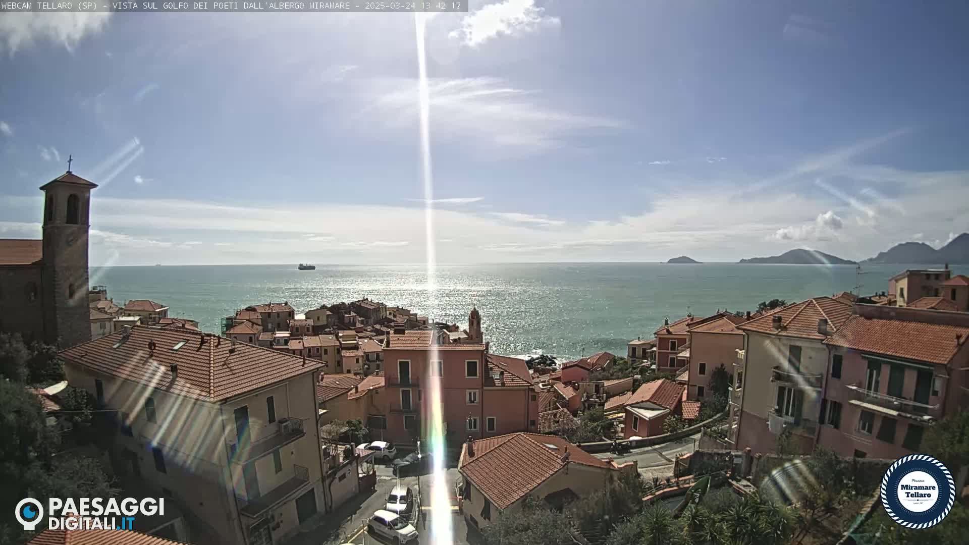 A sunny day overlooking a coastal town with red-tiled roofs and a church steeple, the ocean visible in the background with distant hills and a few ships.