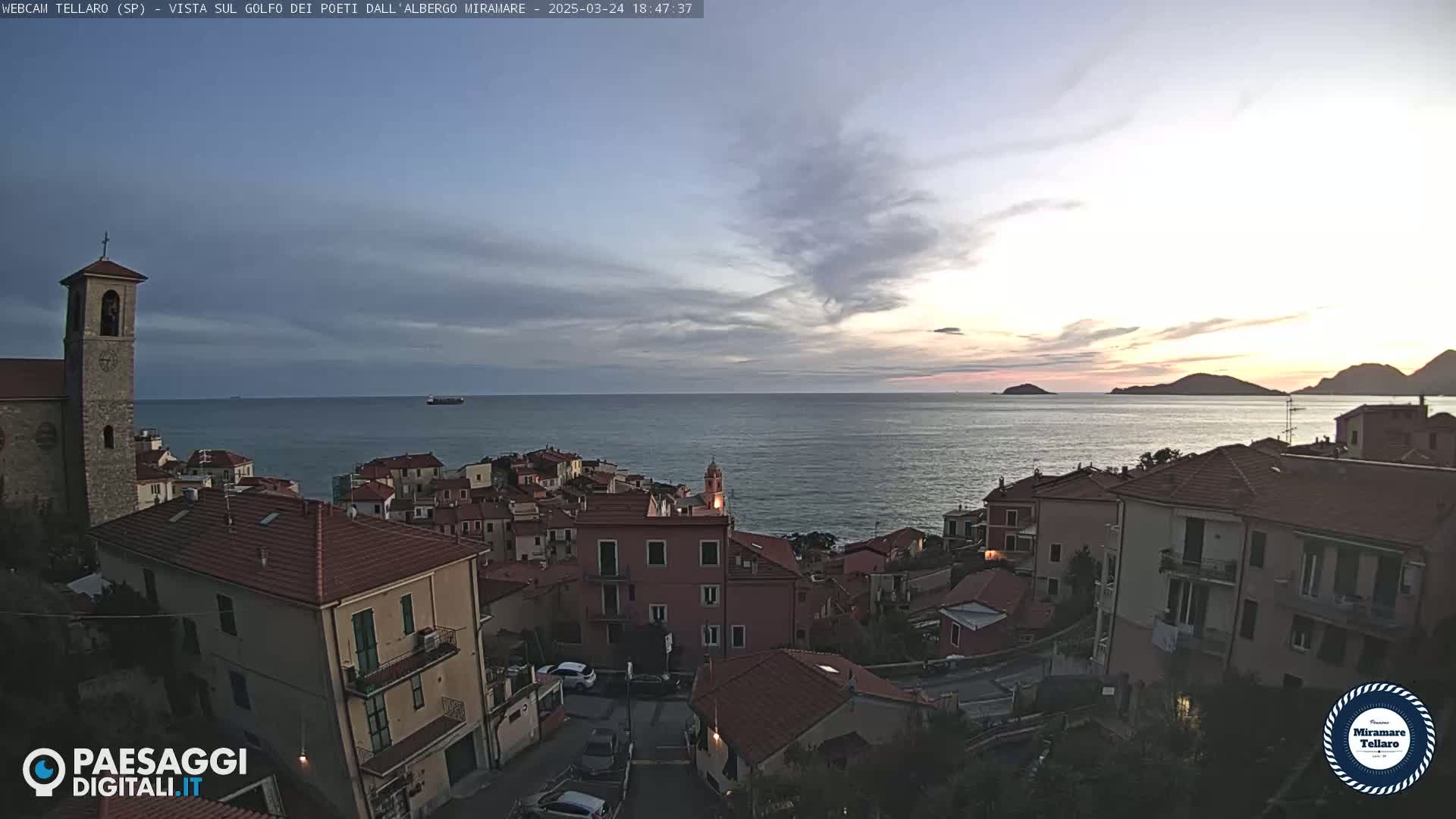A coastal town with red-tiled roofs overlooks a calm sea under a partly cloudy sunset sky.