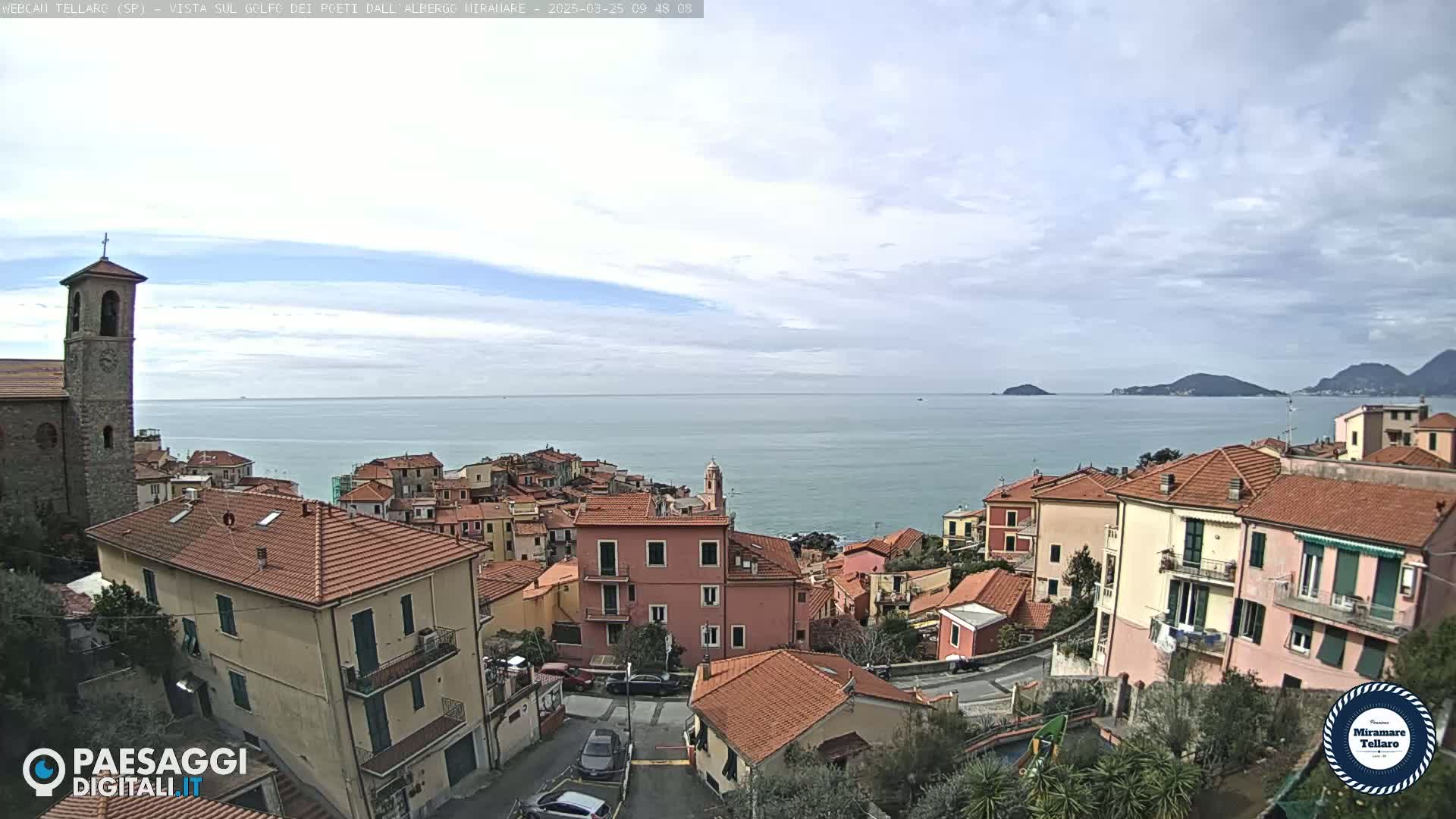 A coastal town with red-tiled roofs overlooks a calm sea under a partly cloudy sky.