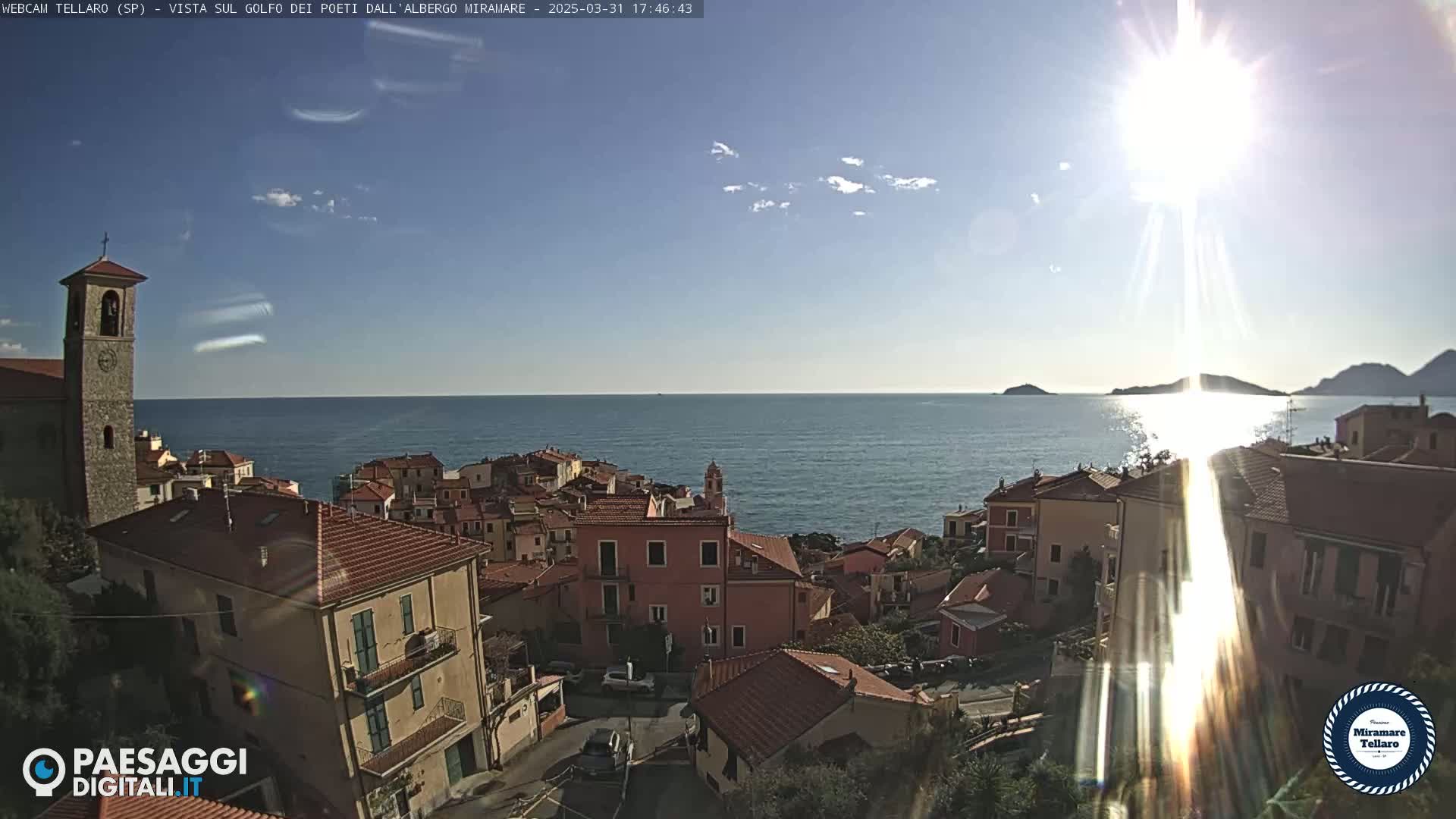 A sunny, coastal town with red-roofed buildings overlooks a calm sea under a mostly clear sky.