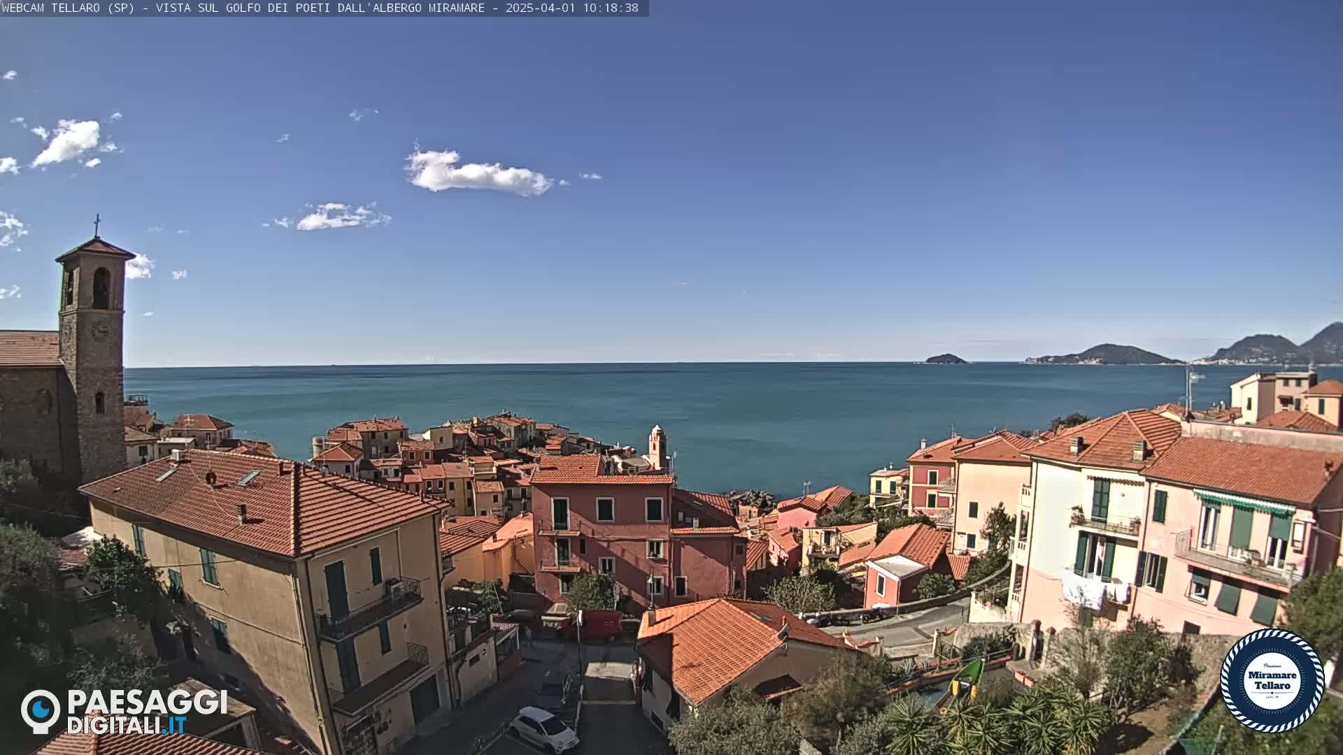 A coastal town with red-tiled roofs and a church steeple overlooks a calm, blue sea under a mostly clear, sunny sky.