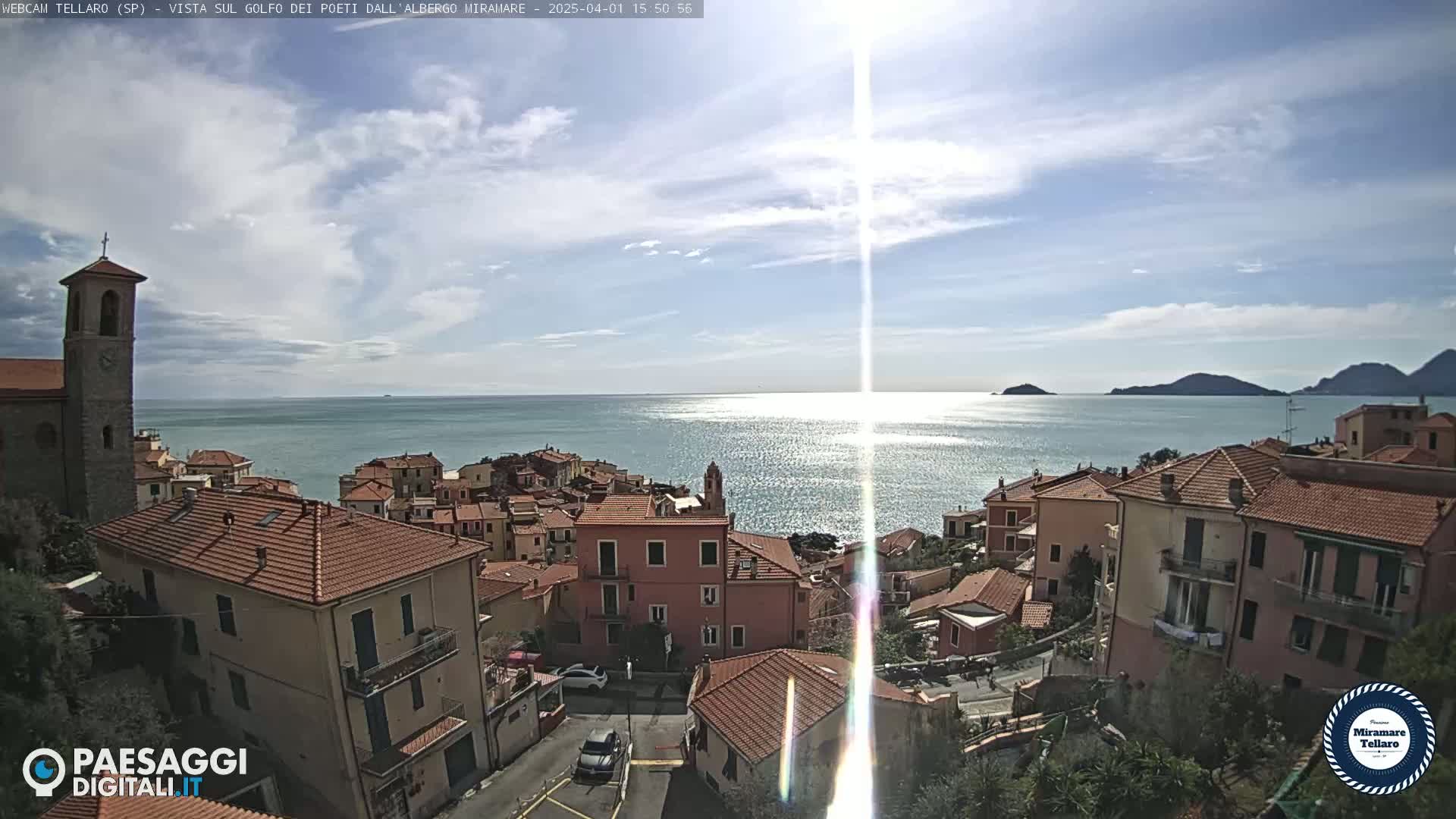 A coastal town with red-tiled roofs sits on a hillside overlooking a calm, sunlit sea under a partly cloudy sky.