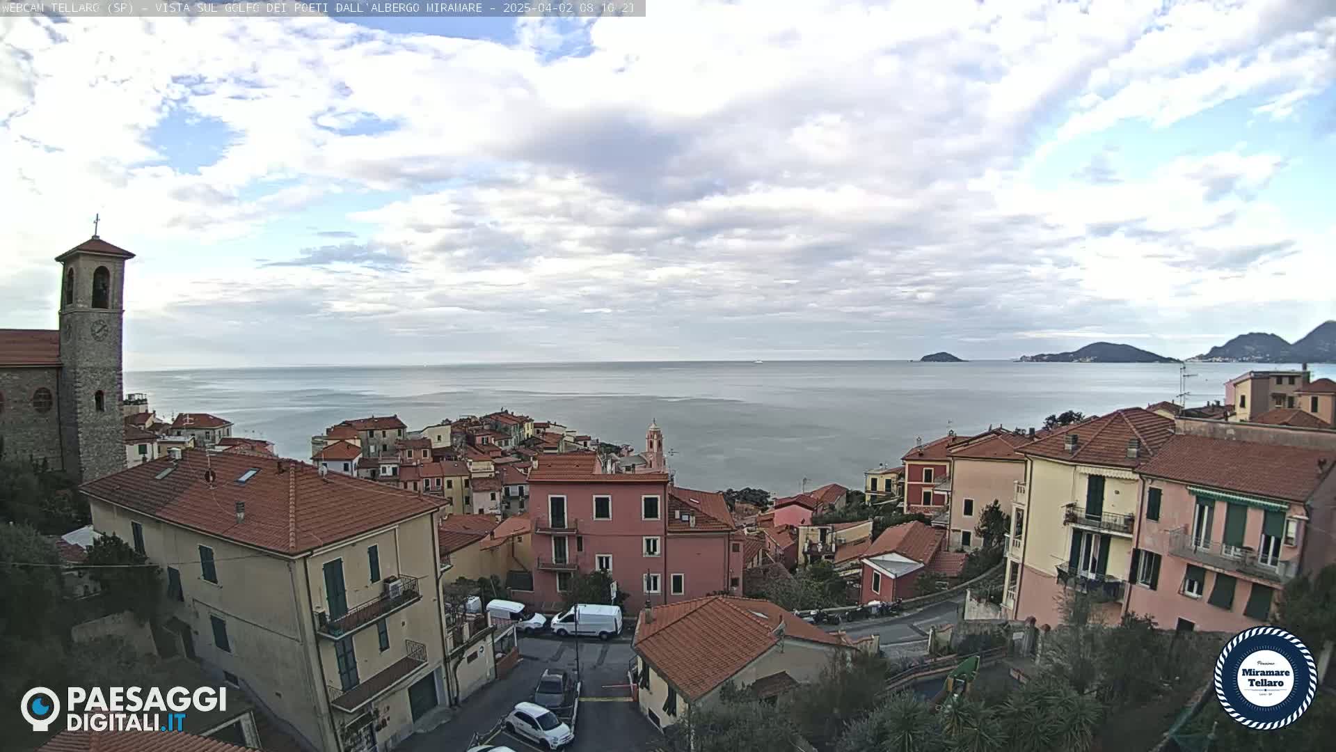 A coastal town with red-tiled roofs and a church bell tower overlooks a calm sea under a partly cloudy sky.