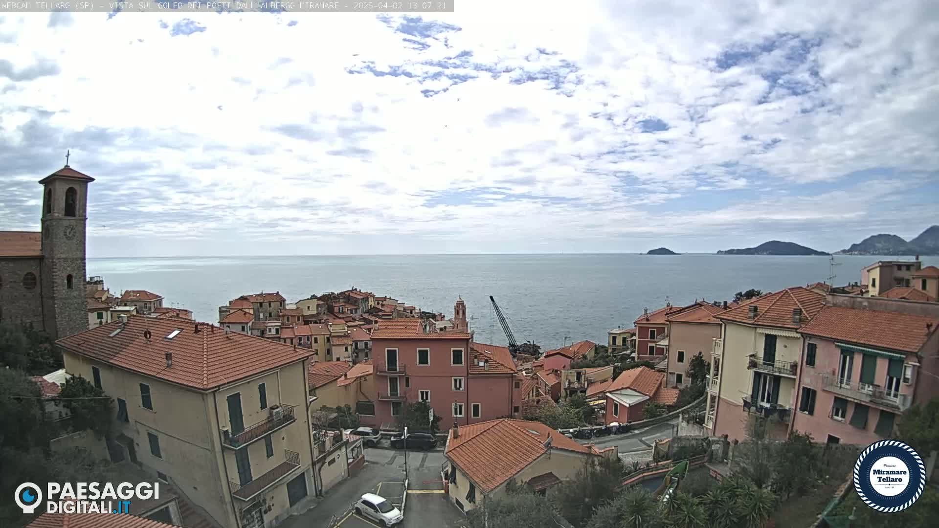 A coastal town with red-roofed buildings overlooks a calm sea under a partly cloudy sky.