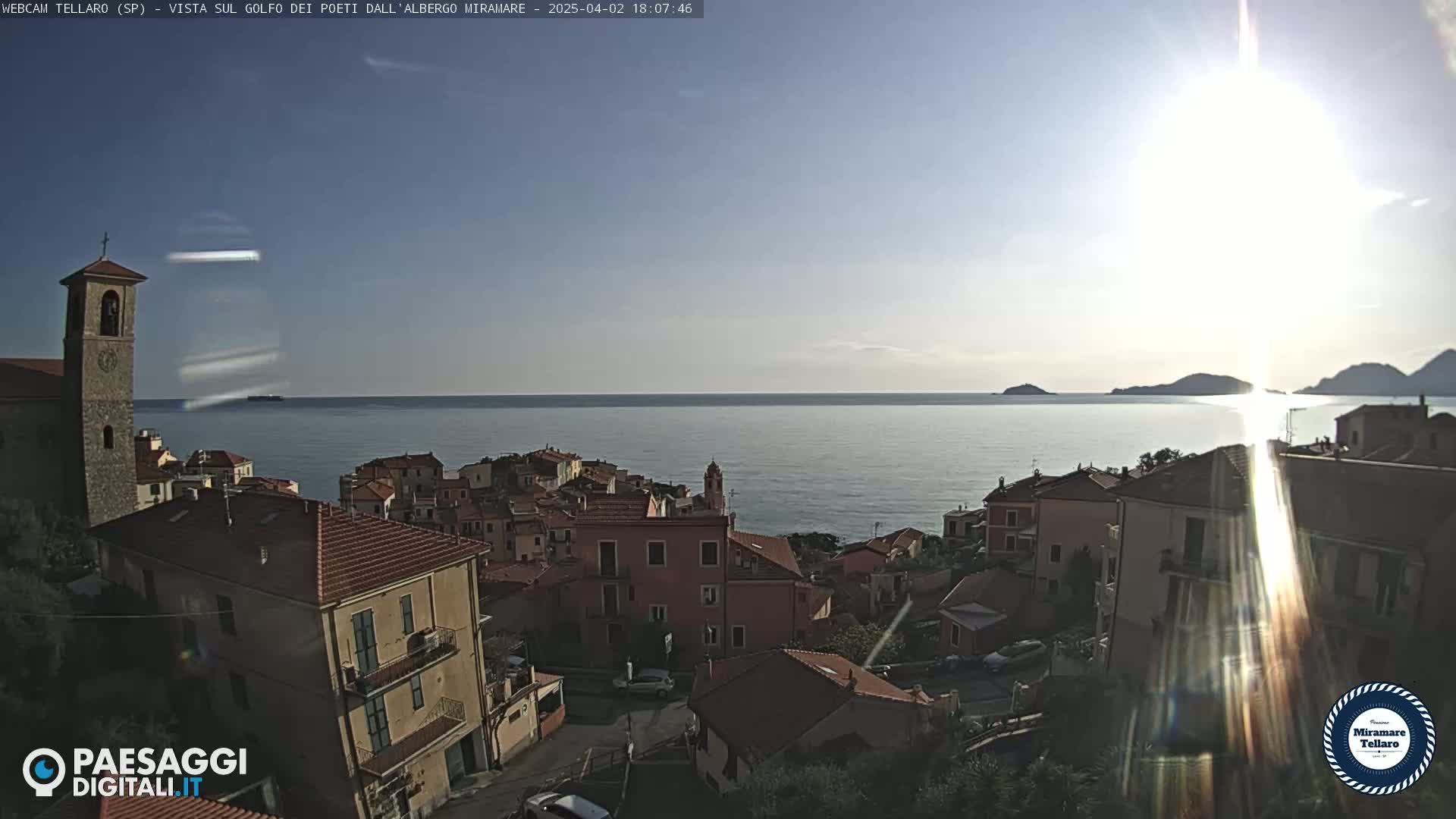 A coastal town with red-tiled roofs and a church steeple overlooks a calm sea under a bright, sunny sky.