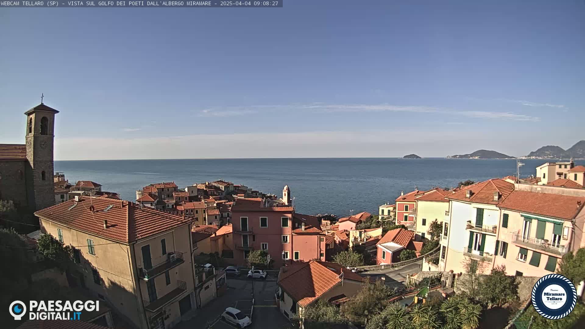 A coastal town with red-tiled roofs and a church steeple overlooks a calm blue sea under a clear sky.
