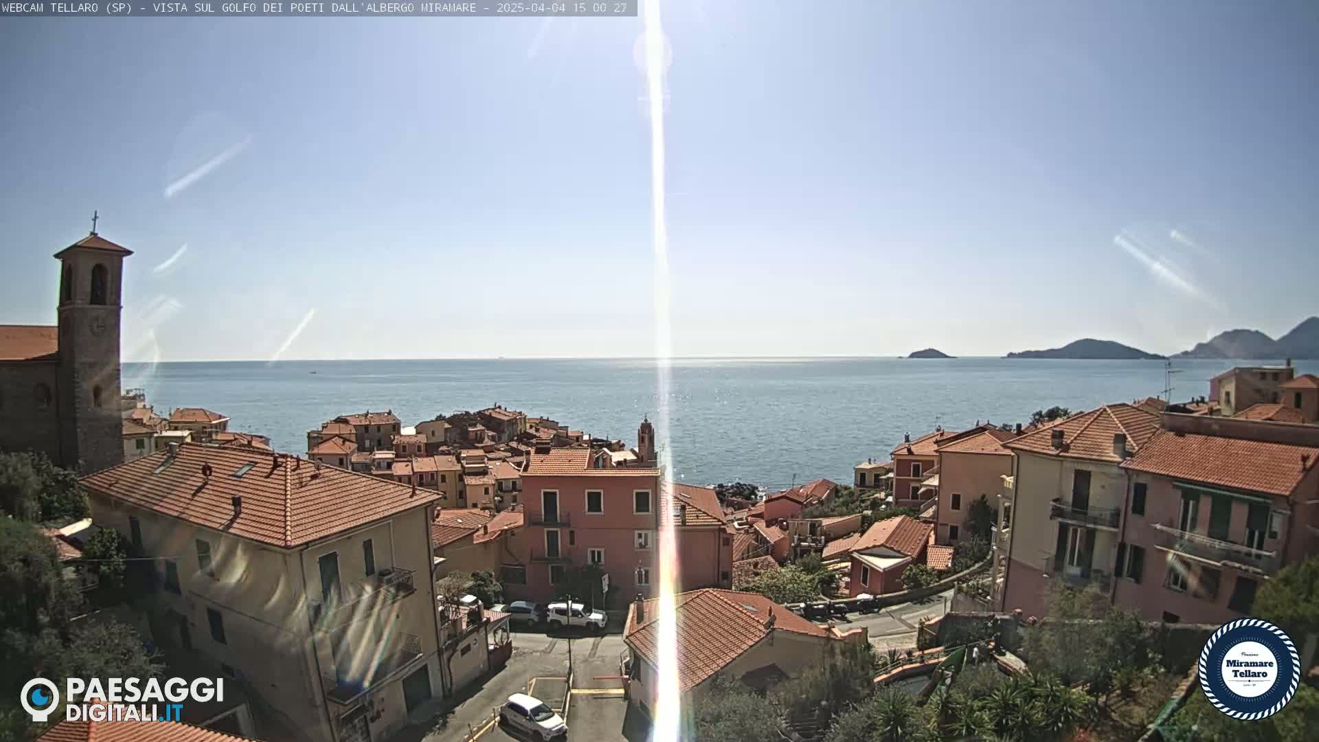 A coastal town with red-tiled roofs and a church steeple overlooks a calm, blue sea under a clear, sunny sky.