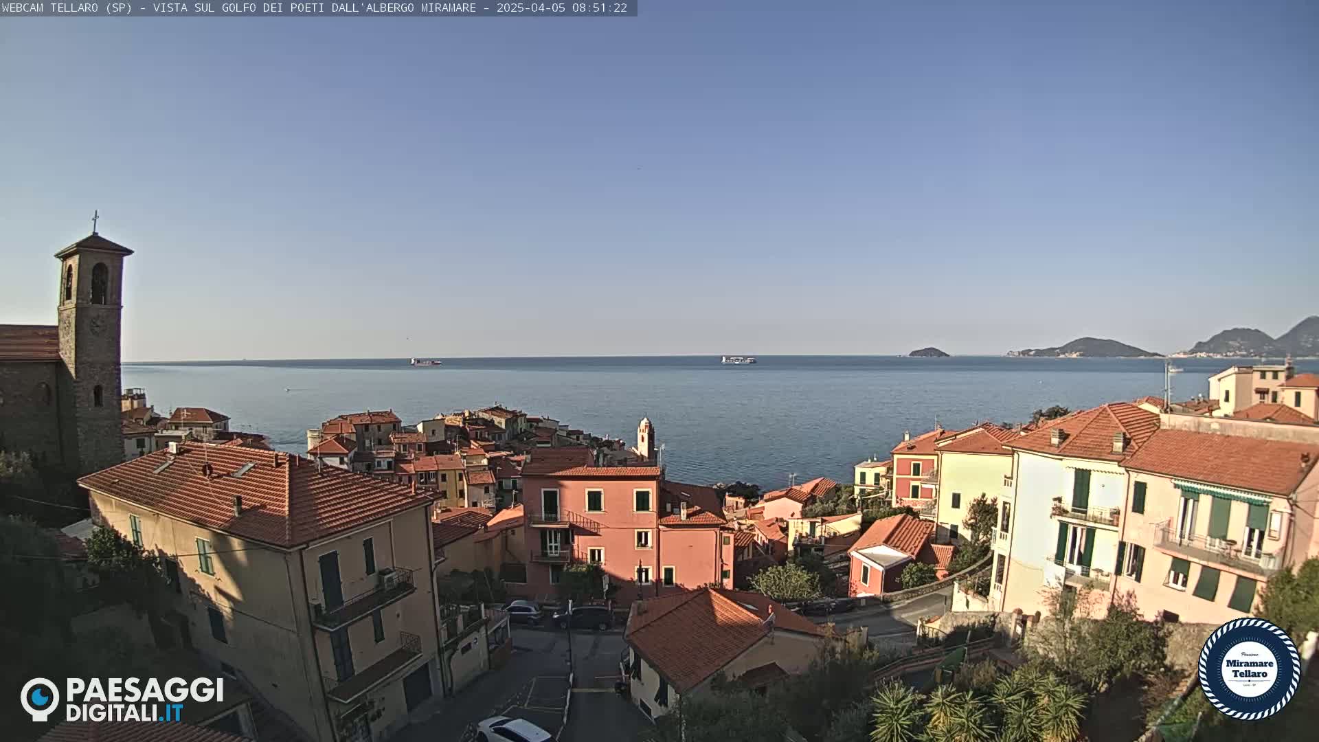 A coastal town with red-roofed buildings overlooks a calm sea under a clear blue sky.