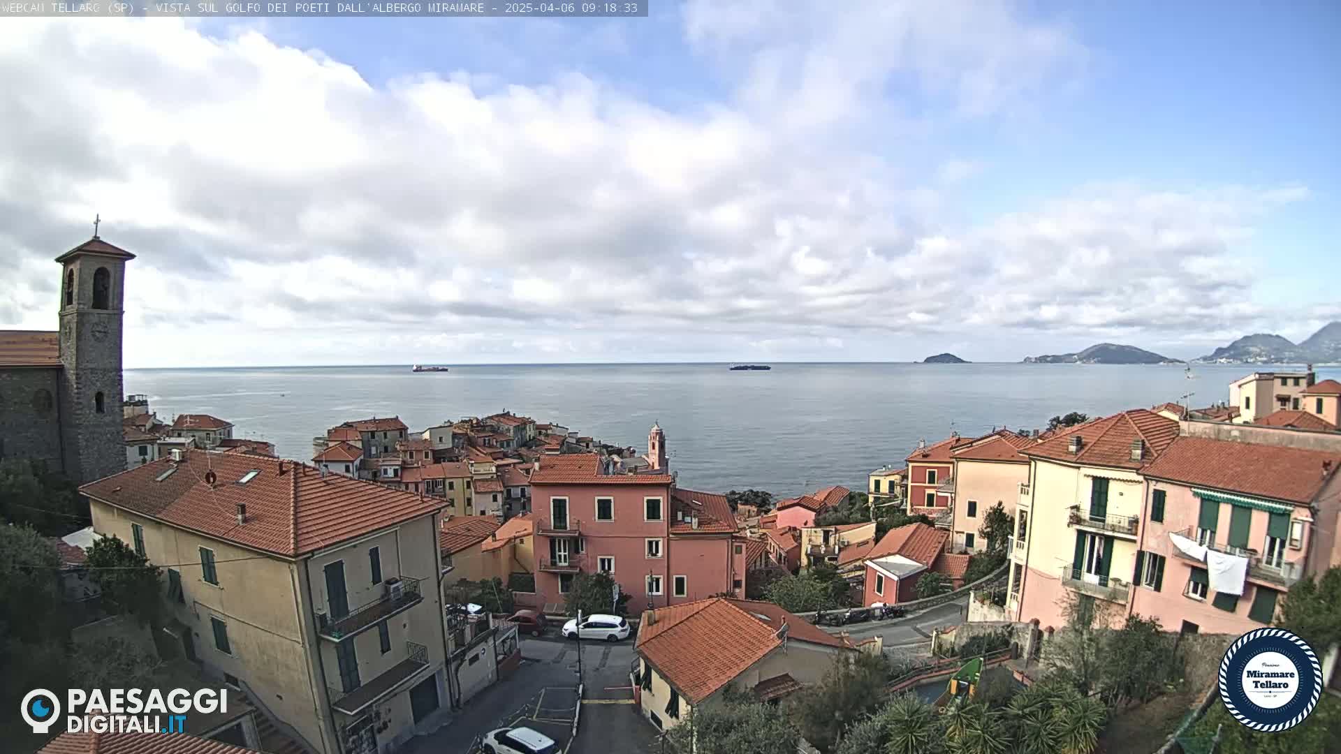 A coastal town with red-tiled roofs overlooks a calm sea under a partly cloudy sky.
