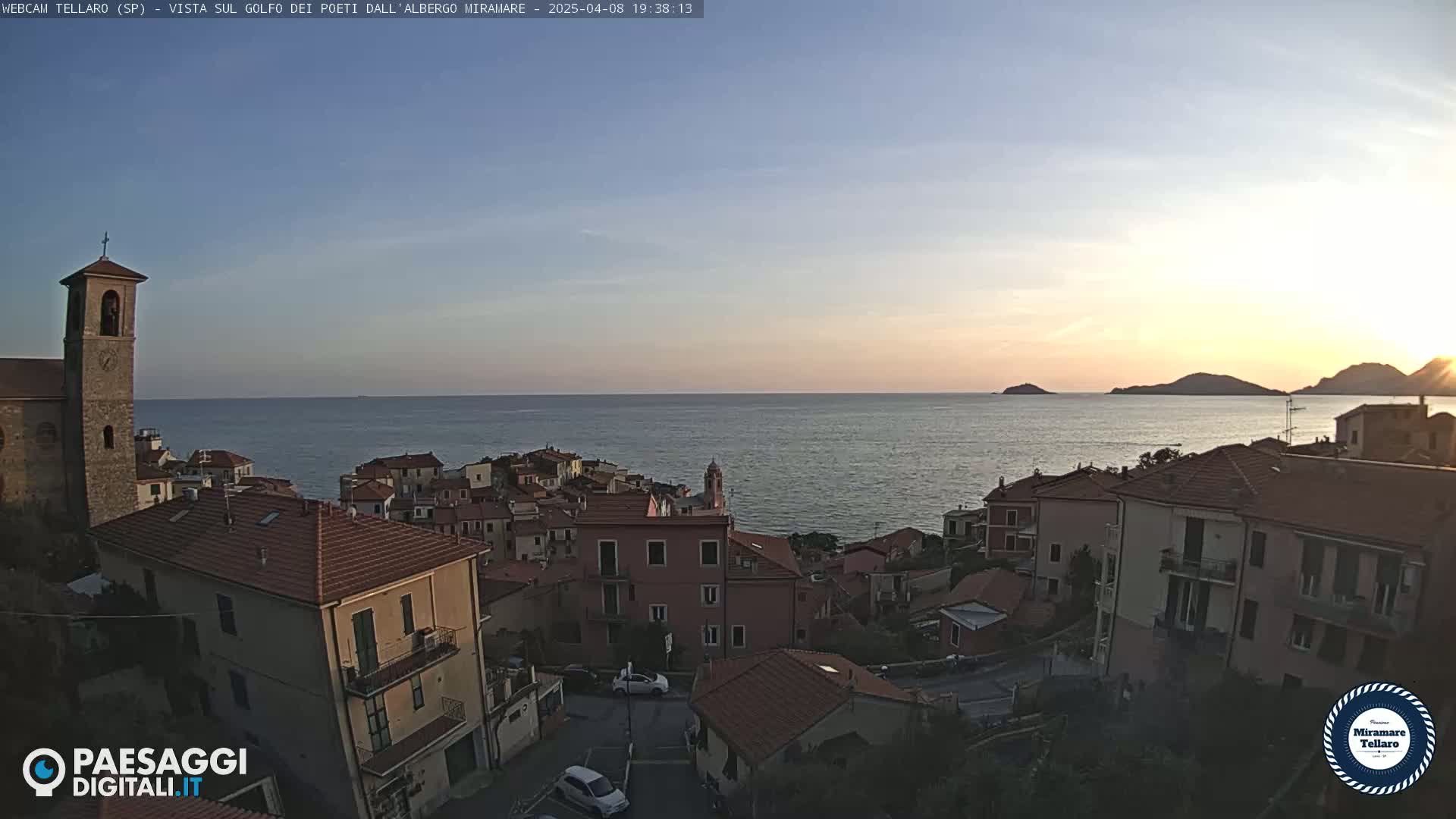 A coastal town with red-tiled roofs overlooks a calm sea at sunset under a clear sky.