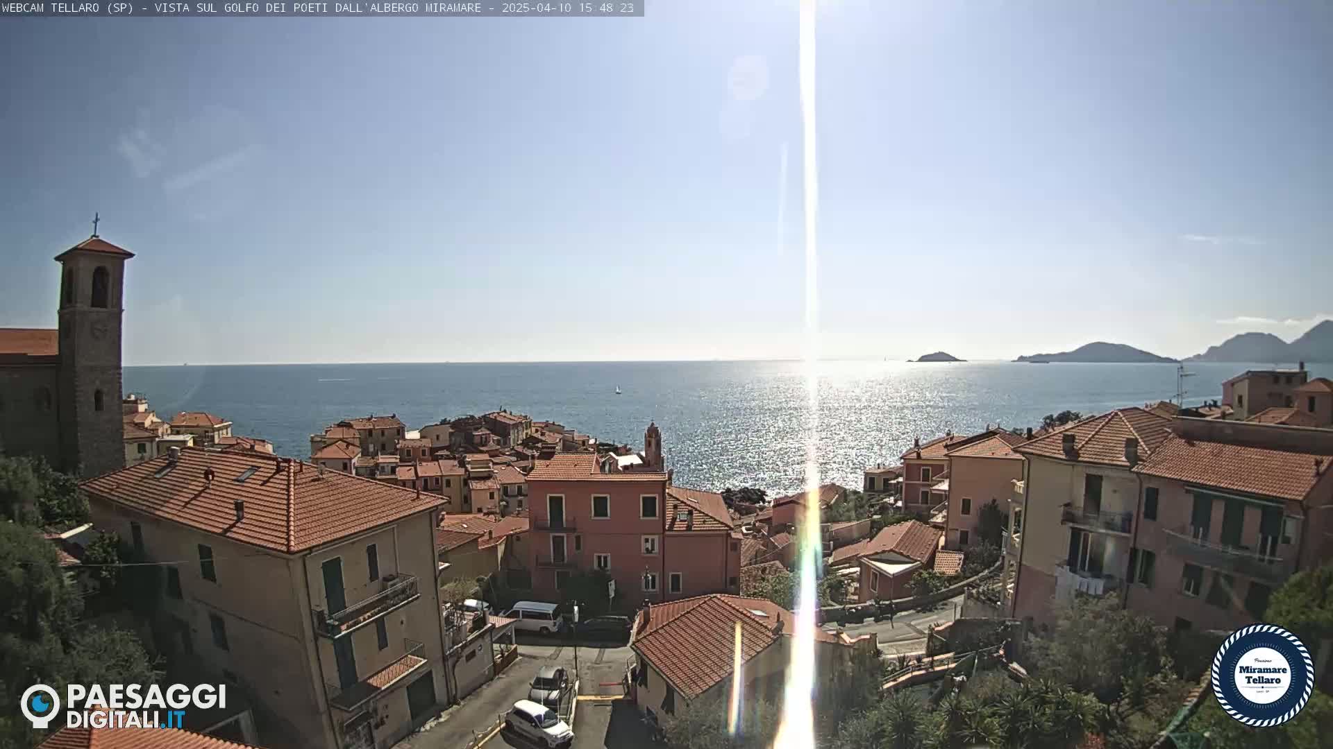 A coastal town with red-tiled rooftops and a church steeple overlooks a calm, sunlit sea under a clear sky.