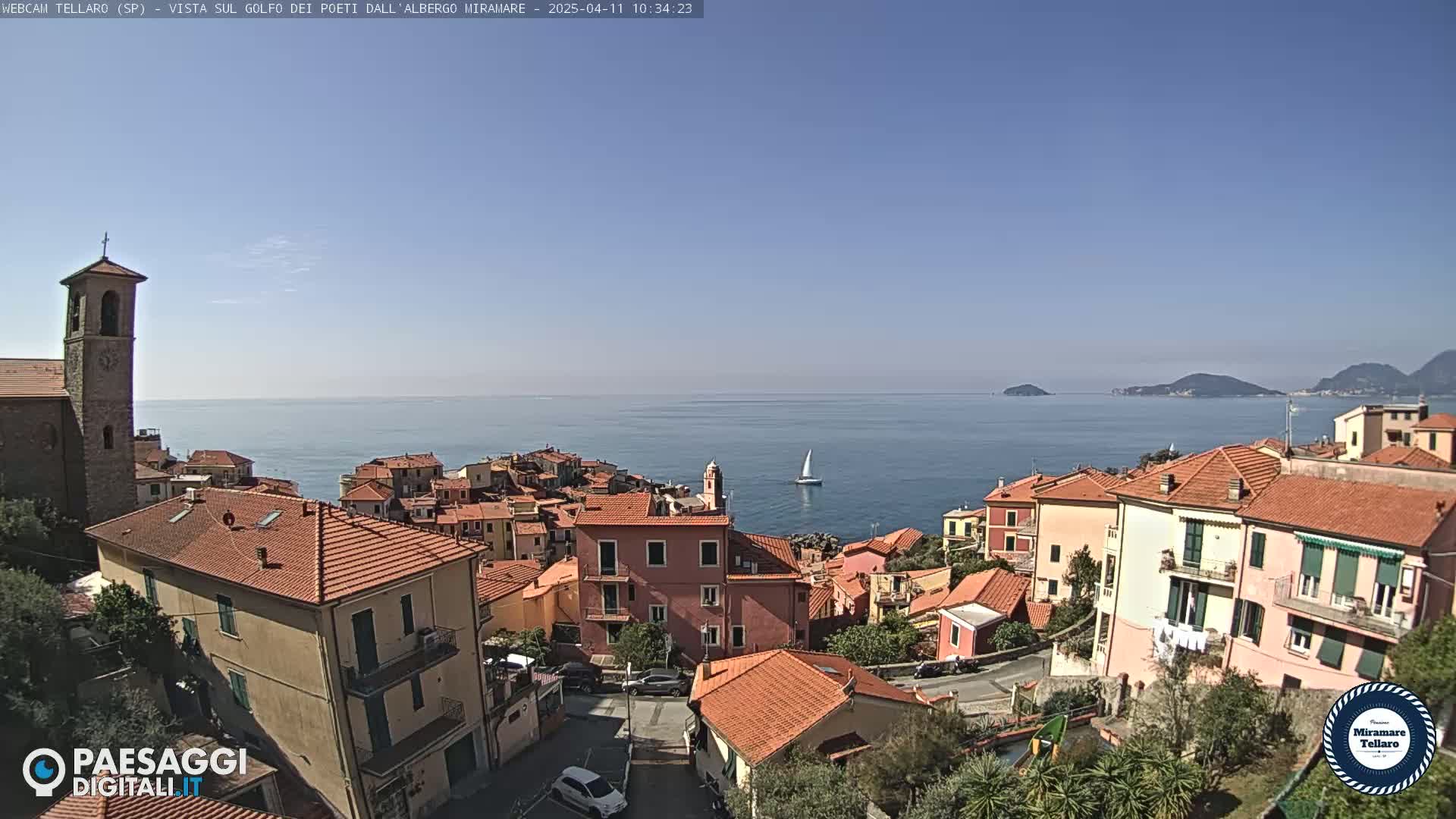 A coastal town with red-tiled roofs and a church steeple overlooks a calm blue sea on a clear day, with a sailboat visible in the distance.