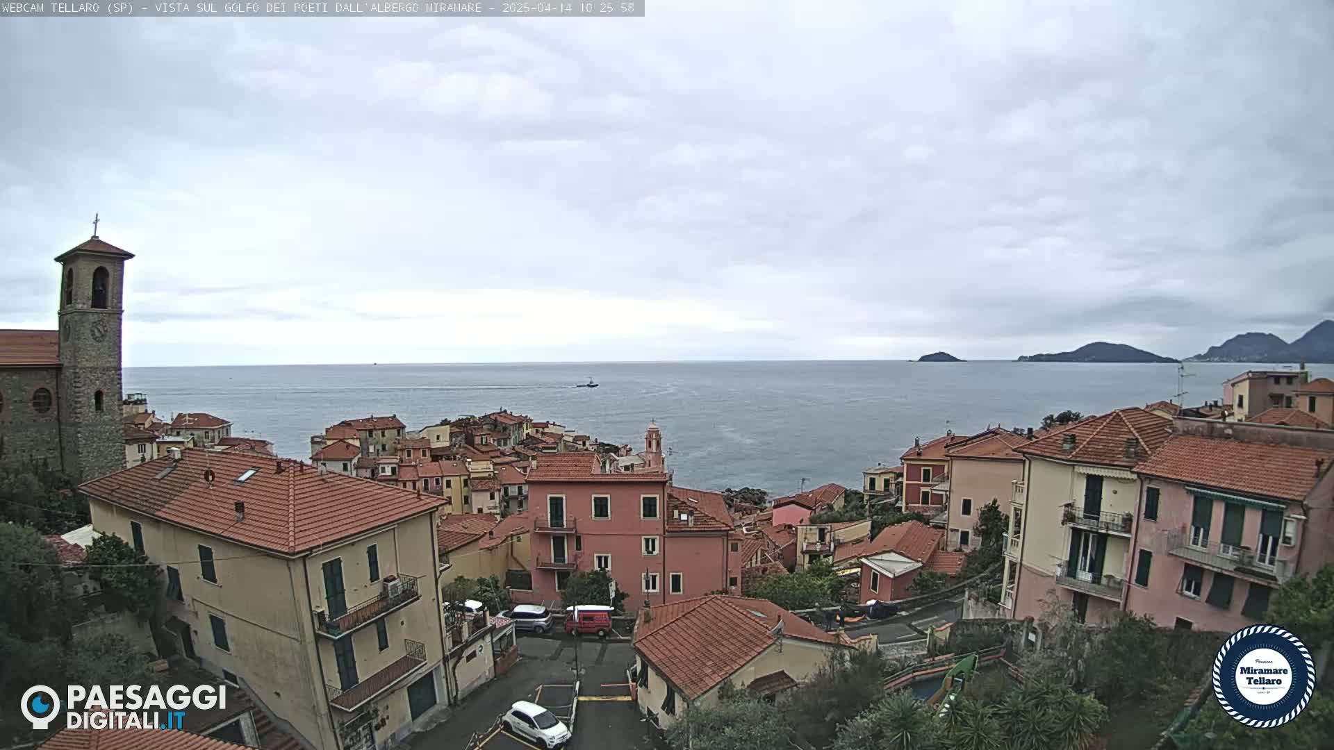 A coastal town with red-tiled roofs and a church bell tower overlooks a calm sea under an overcast sky.