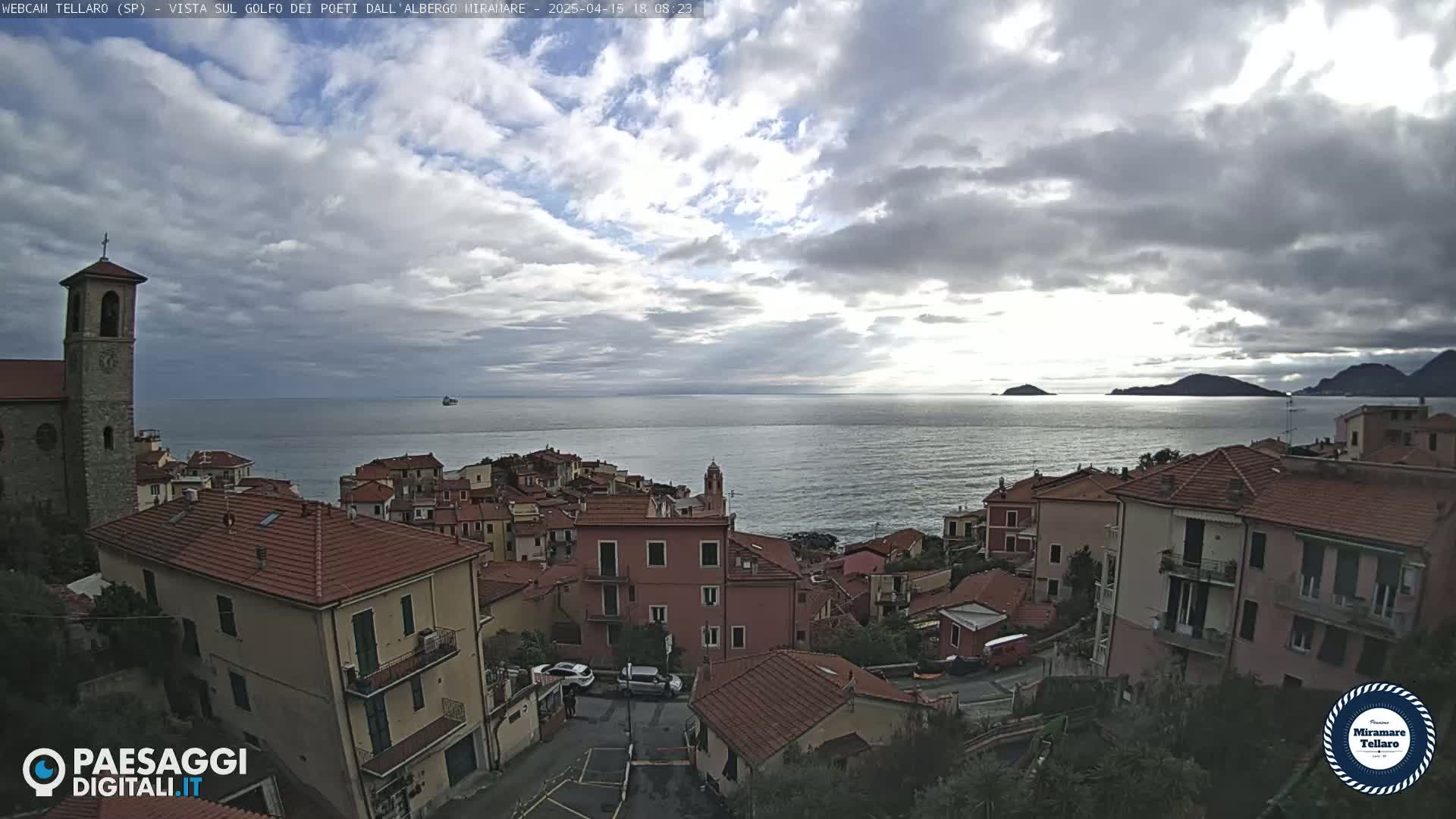 A coastal town with red-tiled roofs overlooks a calm sea under a partly cloudy sky.