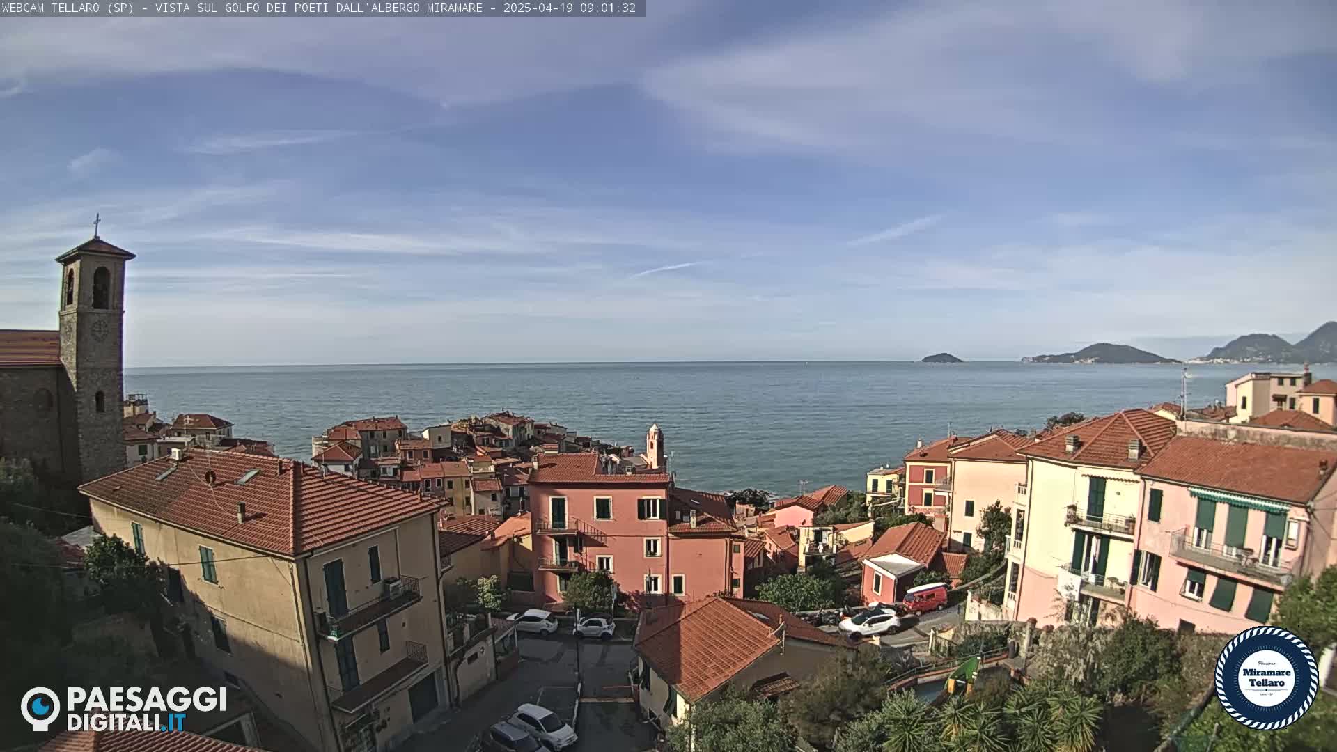 A coastal village with red-tiled roofs overlooks a calm sea under a mostly clear blue sky.
