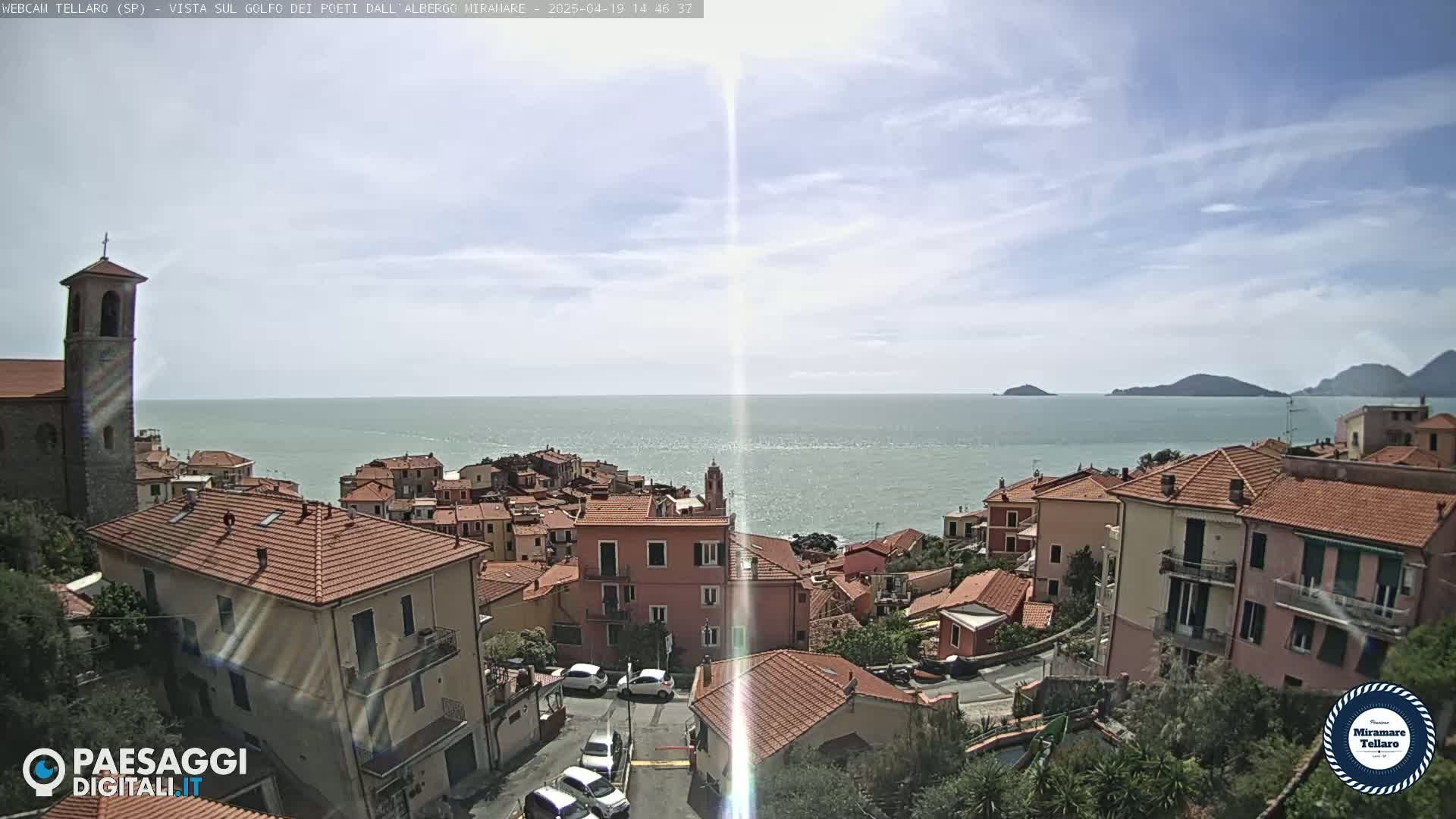 A coastal town with red-tiled roofs and a church steeple overlooks a calm sea under a partly cloudy sky.