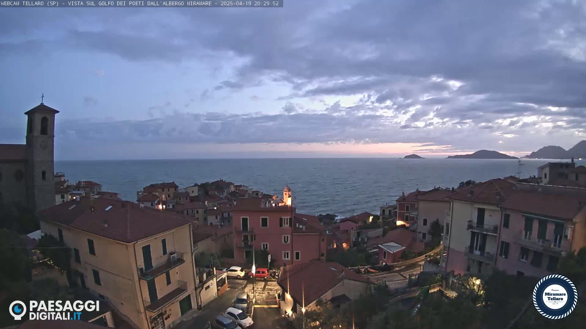 A coastal town with red-roofed buildings sits on a hill overlooking a calm sea under a partly cloudy twilight sky.