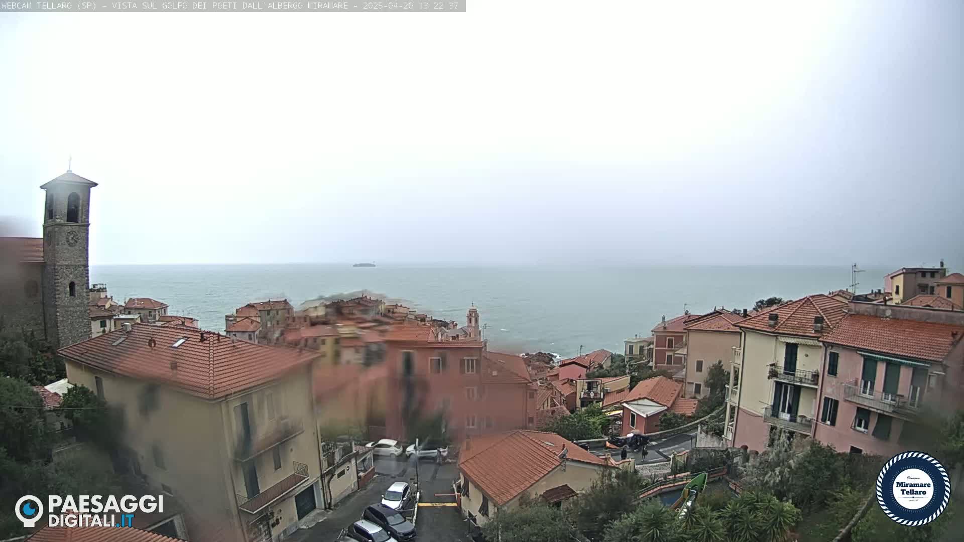 A coastal town with red-tiled roofs and a bell tower overlooks a calm sea under an overcast sky.