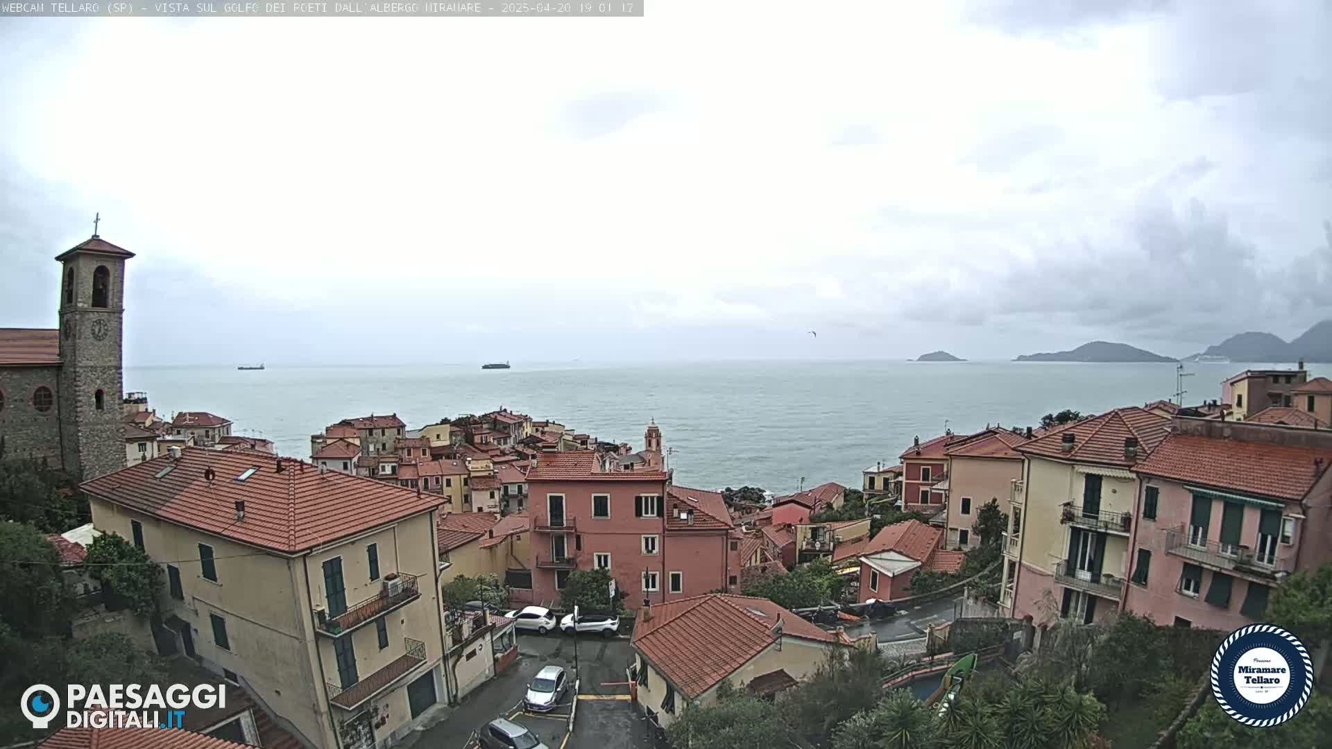 A coastal town with red-tiled roofs and a church, overlooks a calm sea under a cloudy sky.