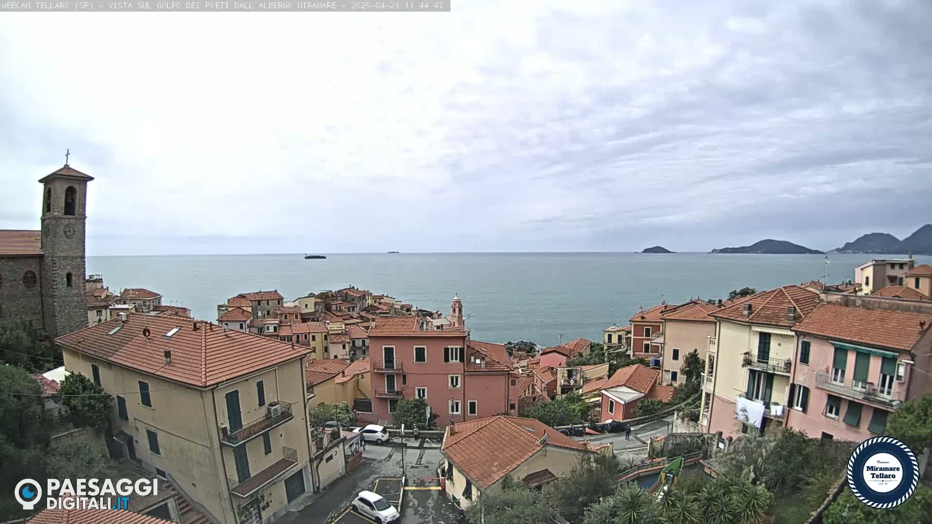 Overlooking a coastal town with red-tiled roofs and a church, a calm sea stretches to distant hills under a cloudy sky.