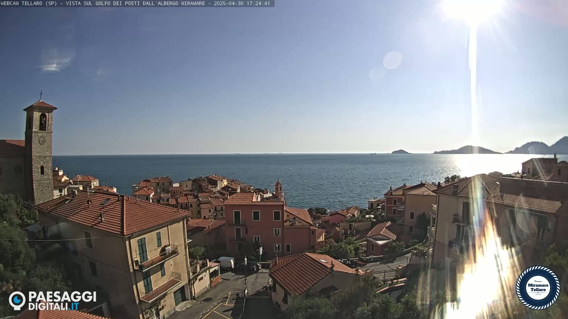 A coastal town with red-tiled roofs nestles beside a calm sea under a bright, sunny sky.