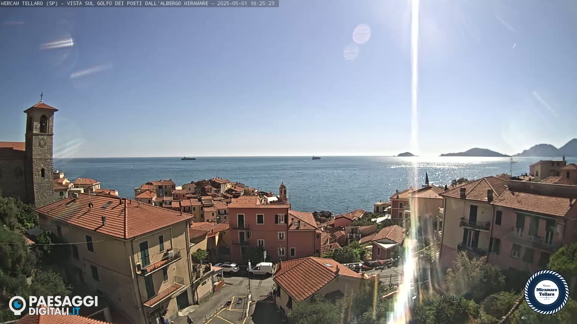 A sunny coastal town with red-tiled roofs overlooks a calm blue sea and distant islands.