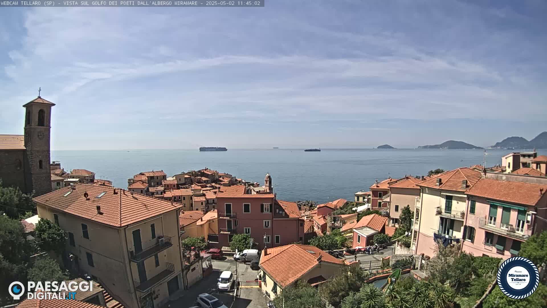 A coastal town with red-roofed buildings overlooks a calm sea under a mostly sunny sky, with several ships visible on the horizon.