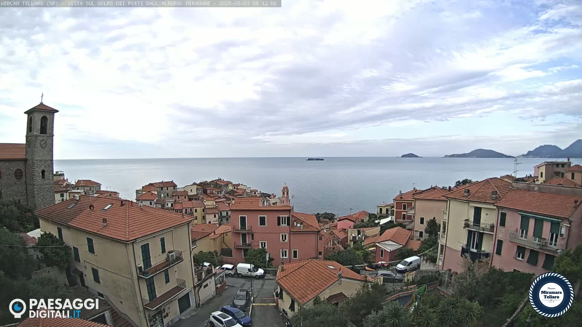 A coastal town with red-tiled roofs and a church steeple overlooks a calm sea under a partly cloudy sky.