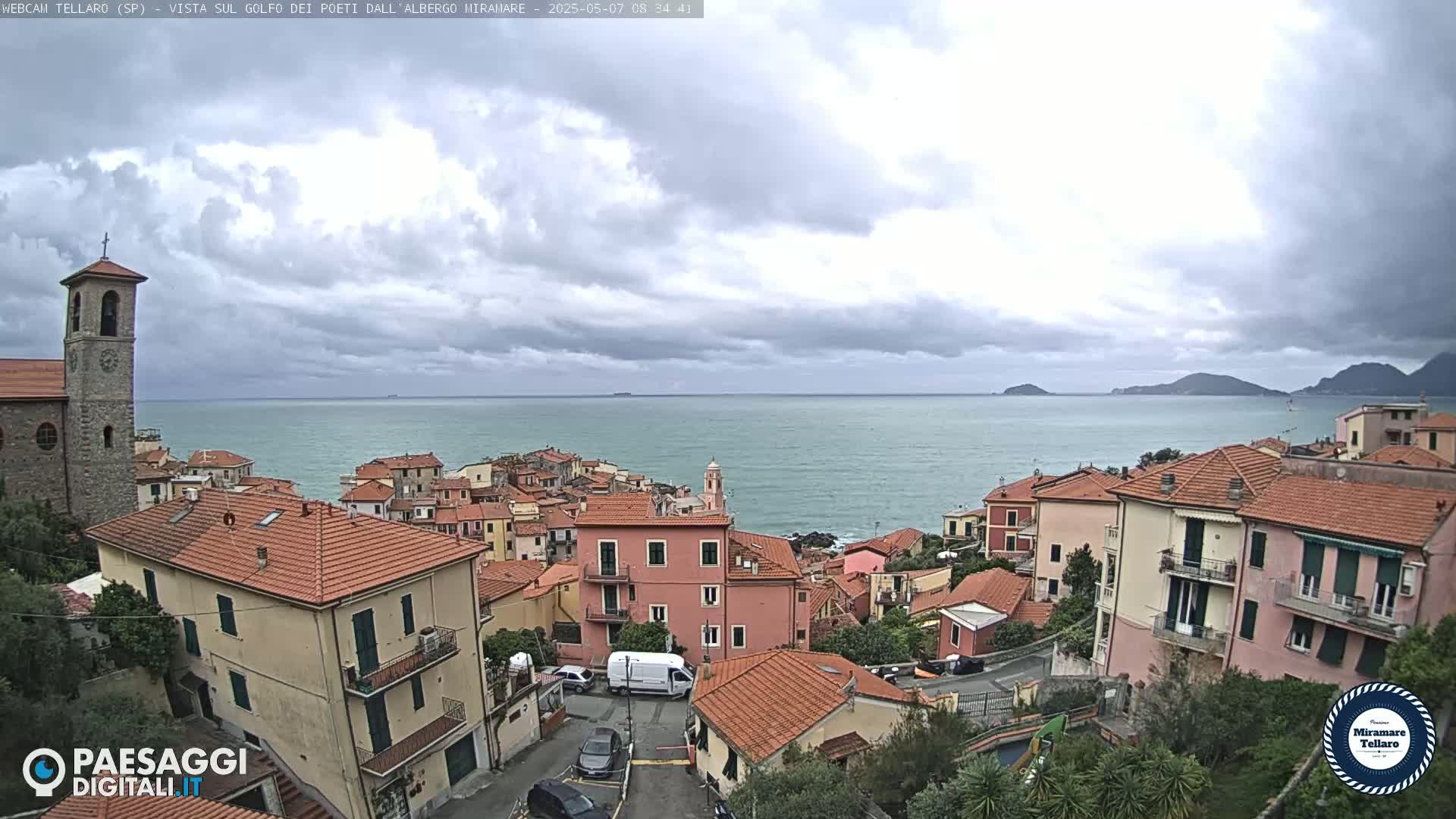 A coastal town with red-tiled roofs and a church steeple overlooks a calm sea under a cloudy sky.