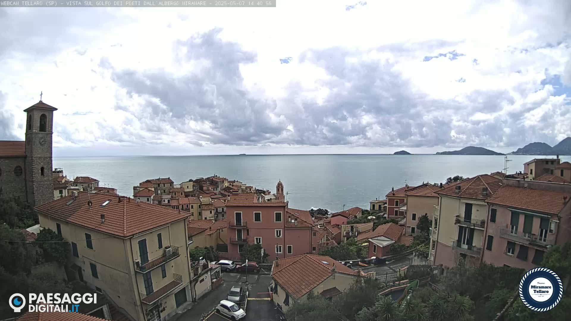 A coastal town with red-tiled roofs overlooks a calm sea under a partly cloudy sky.