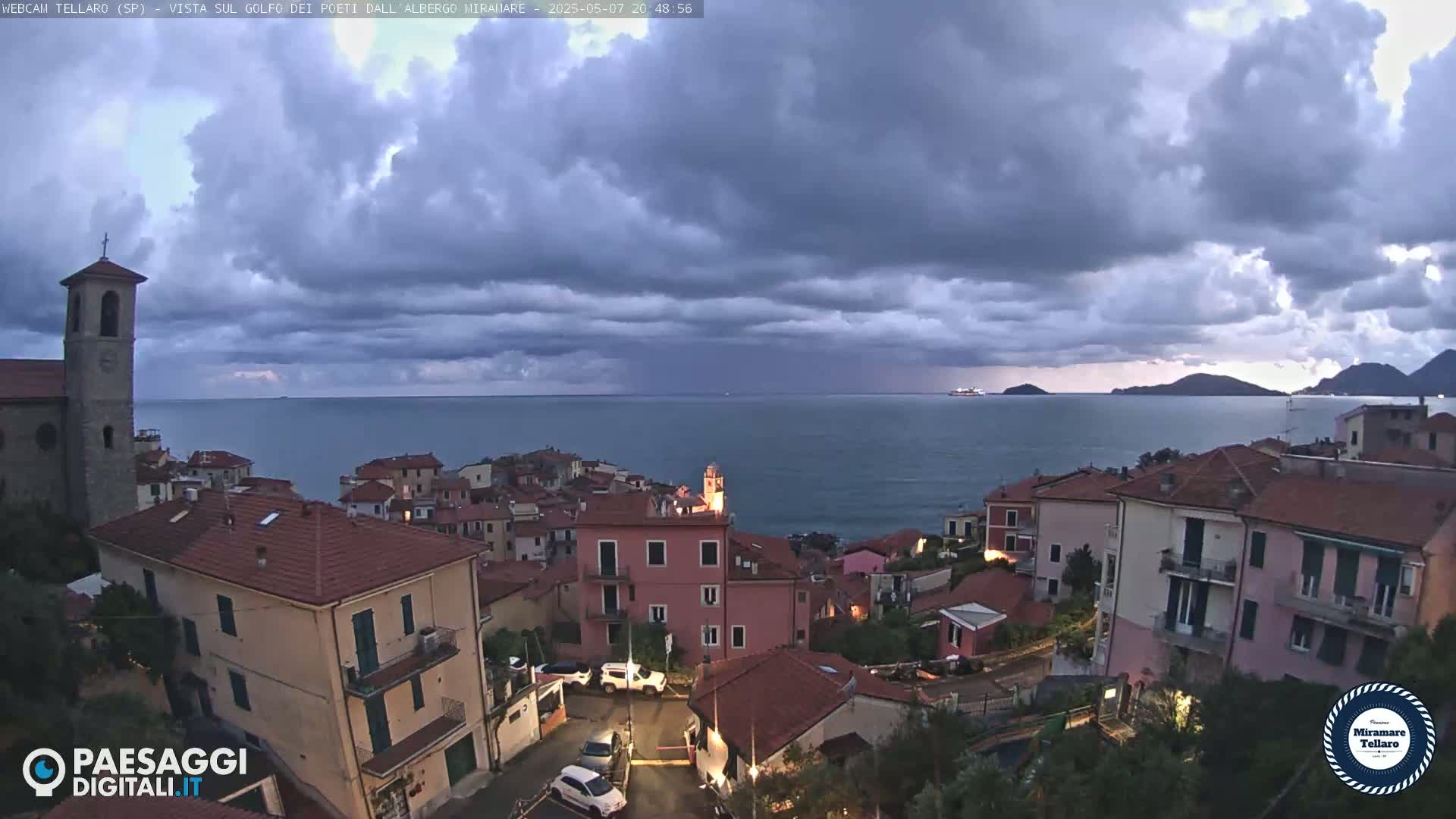 A coastal town with red-tiled roofs sits under a partly cloudy, twilight sky overlooking a calm sea with distant hills and a ship.