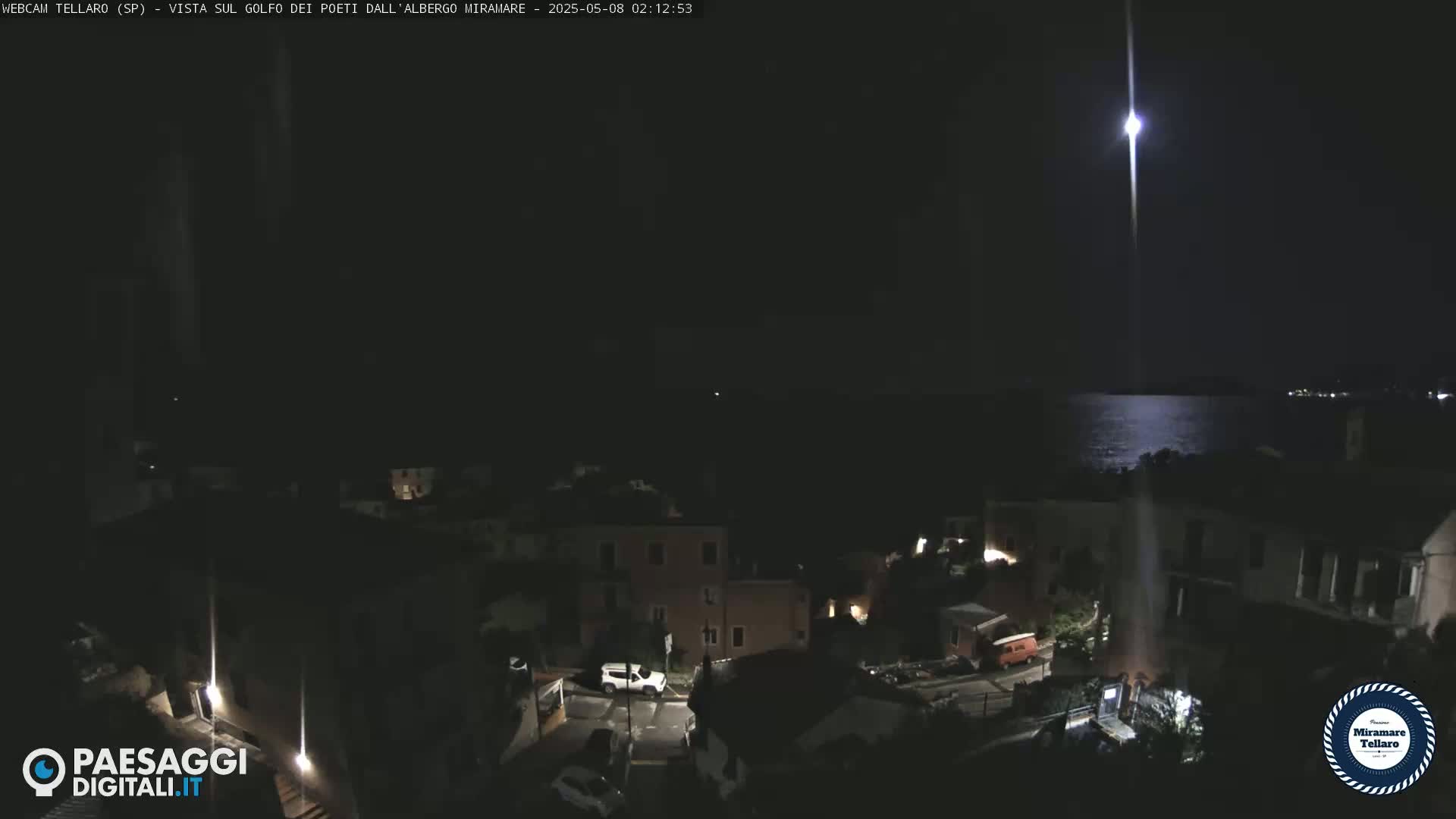 A nighttime view of a coastal town, illuminated by streetlights, overlooks a calm sea reflecting a bright moon.