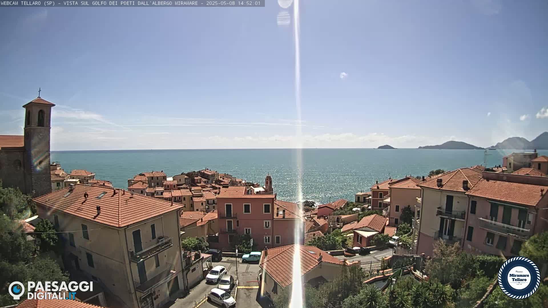 A sunny coastal town with red-tiled roofs overlooks a calm, blue sea under a mostly clear sky.