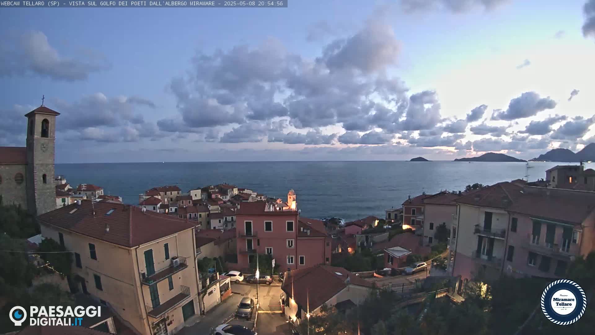 A coastal town with red-roofed buildings overlooks a calm sea under a partly cloudy twilight sky.