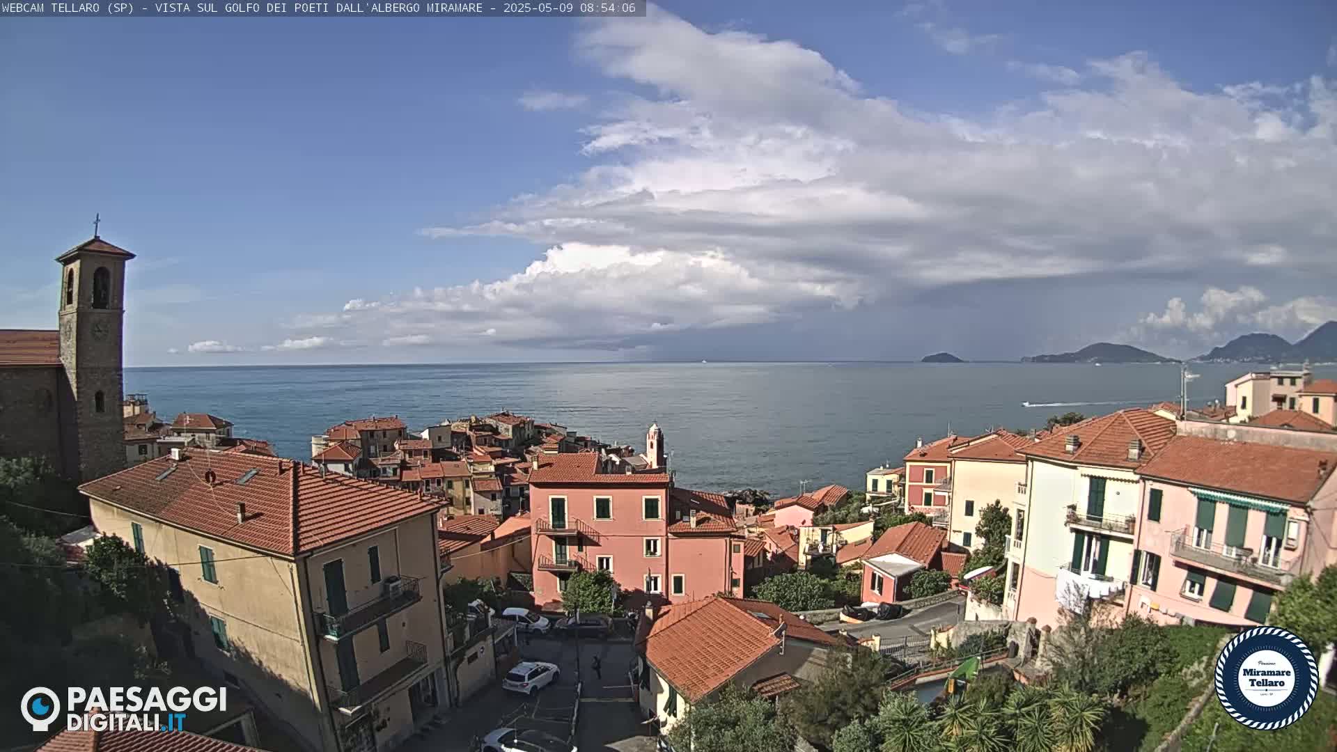 A coastal village with red-tiled roofs and a church steeple overlooks a calm sea under a partly cloudy sky.