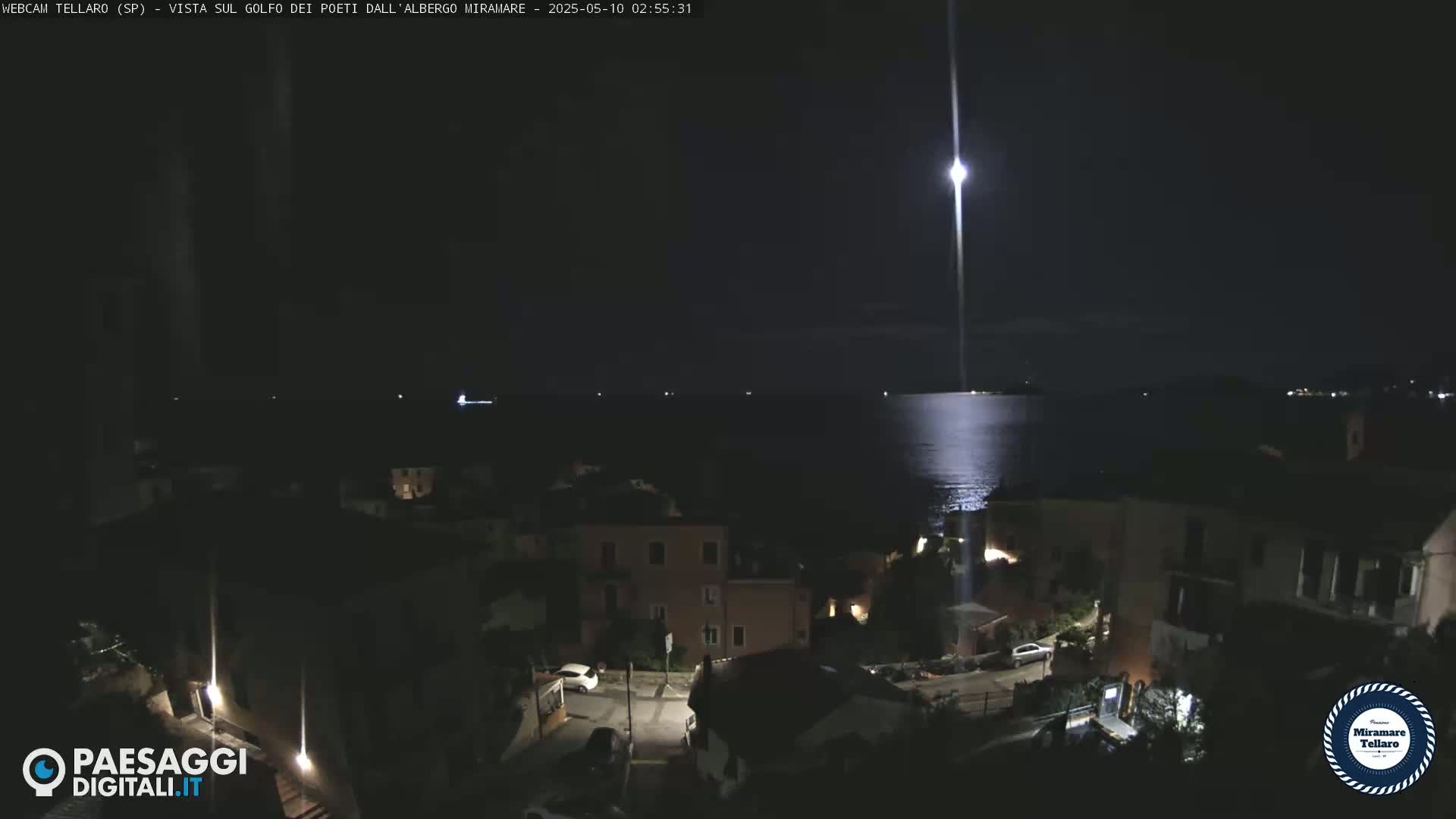 A bright light shines over a dark coastal town at night, illuminating a calm body of water.