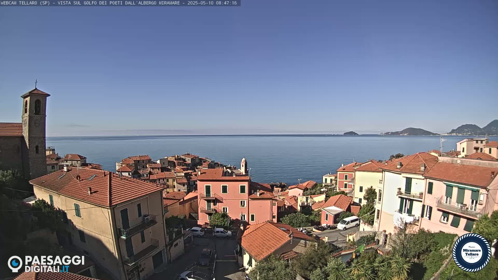 A coastal town with red-tiled roofs and a church steeple overlooks a calm, blue sea under a clear sky.